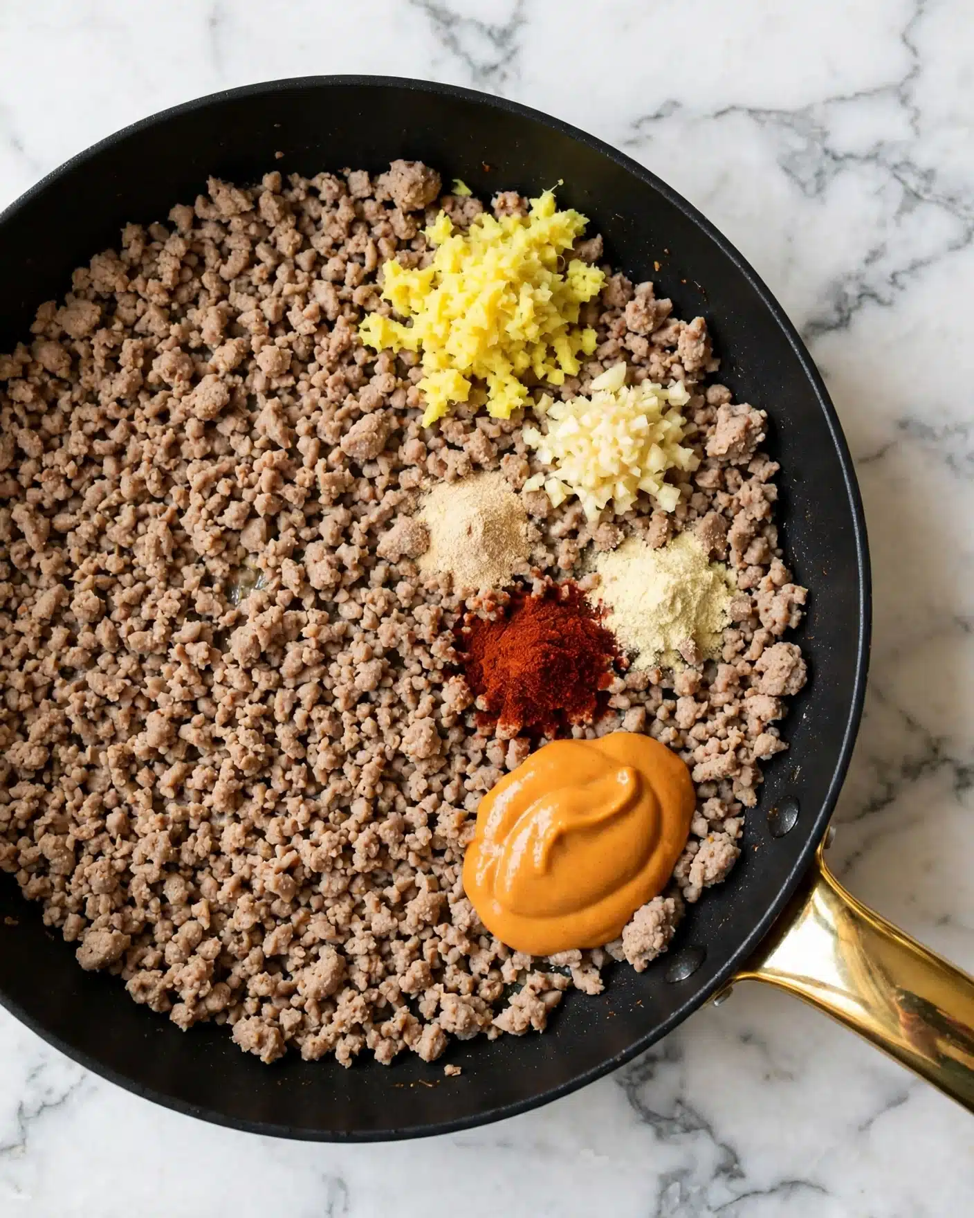 Ground Turkey Rice Bowls with Bang Bang Sauce Recipe 4 The image shows a black pan filled with light brown cooked ground meat, spread evenly as the base layer. On top of the meat, there are five small piles of colorful ingredients: a bright yellow minced layer that looks like fresh ginger at the upper right, a slightly paler minced layer below it, a dark red powder near the center right, a light beige powder above the red, and a smooth, thick orange sauce at the lower right side. The black pan has a shiny gold handle, and the pan is placed on a white marbled surface photo taken with an iphone --ar 4:5 --v 7