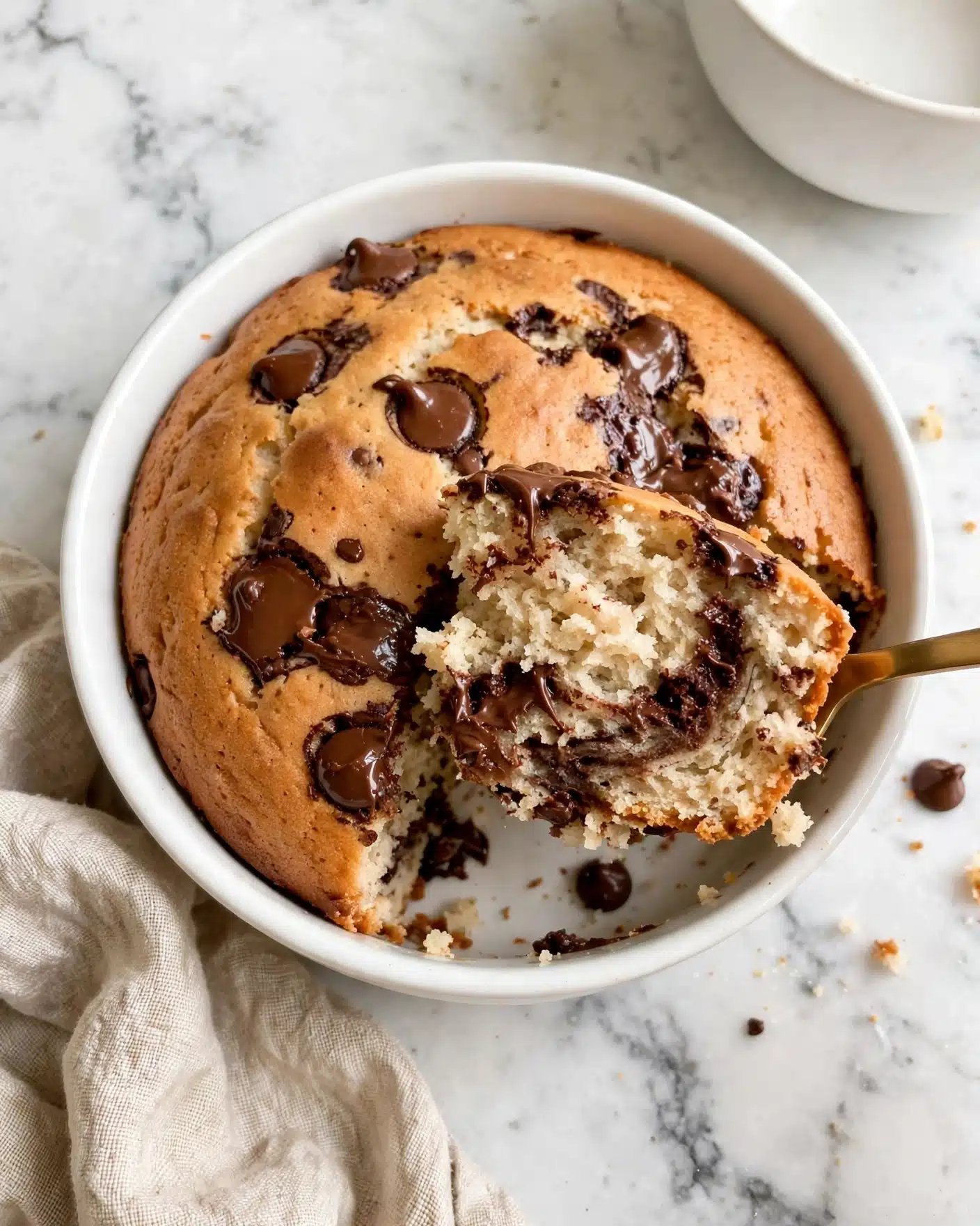 A white round bowl holds a warm, soft cake with a light brown top layer that has chocolate chips scattered on the surface, melted slightly in some spots. The inside of the cake is fluffy and pale beige with swirls of darker melted chocolate throughout. A gold spoon lifts a portion of the cake, showing the moist texture inside. The bowl is placed on a white marbled surface next to a beige textured cloth and a white bowl is partially visible at the top right. Photo taken with an iphone --ar 4:5 --v 7