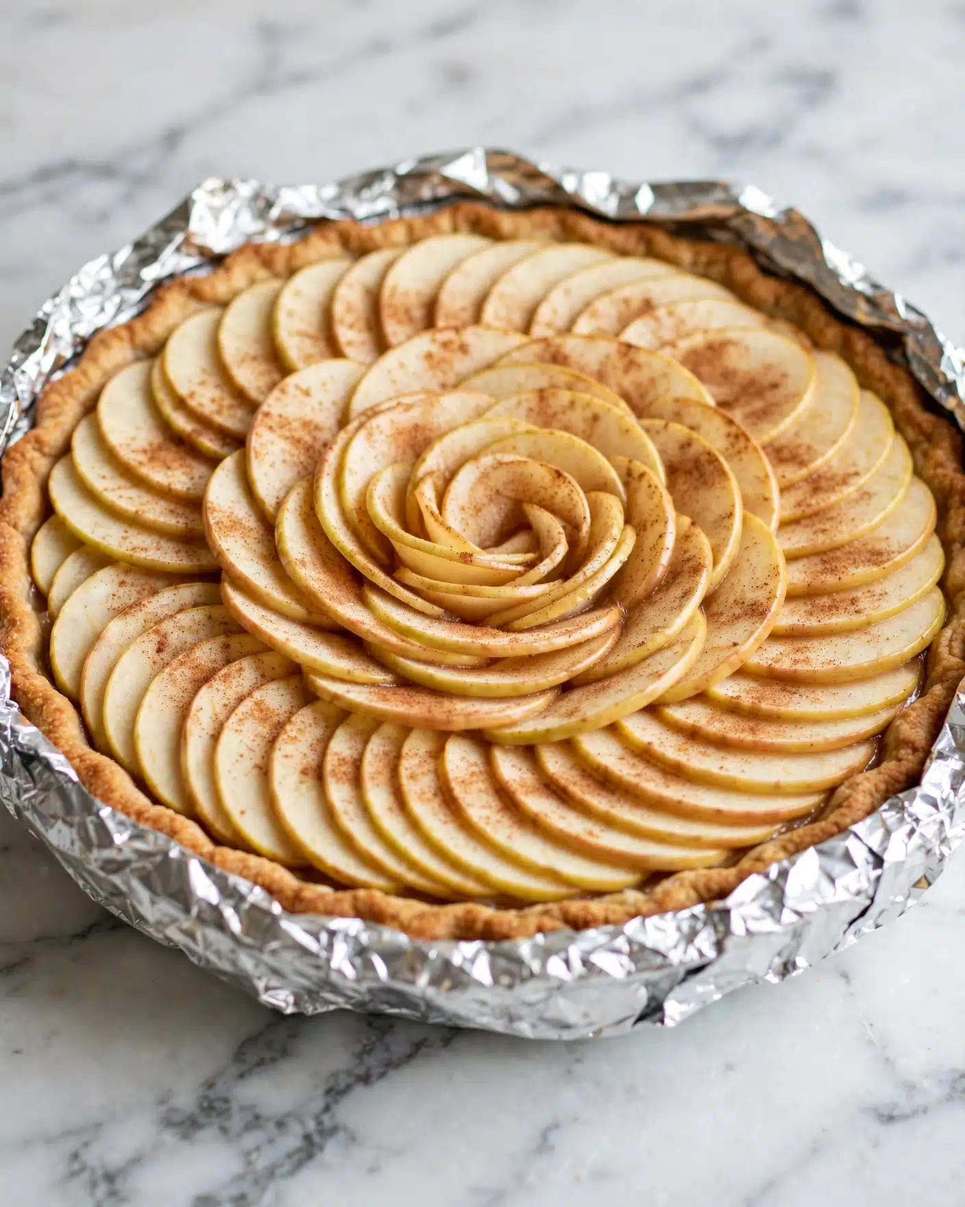 Apple Crisp Cheesecake with Caramel Drizzle Recipe 4 A round pie with a golden brown crust wrapped in foil sits on a white marbled surface. Inside, there are thinly sliced apple pieces arranged in overlapping circular layers covering the entire pie. The apples are light yellow with a dusting of cinnamon, giving them a warm, speckled light brown color. The layers are clearly visible with the apples gently curved, neatly packed from the edge to the middle of the pie. Photo taken with an iphone --ar 4:5 --v 7