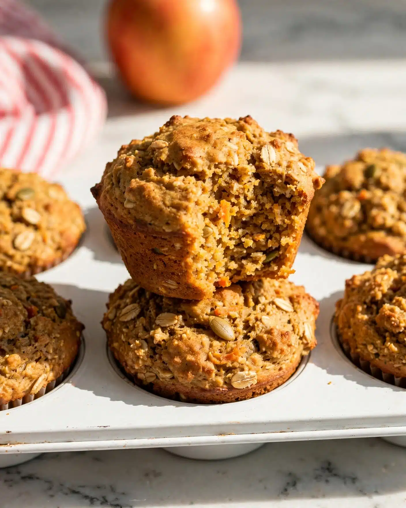 Healthy Pumpkin Carrot Apple Muffins Recipe 5 The image shows a close-up of two golden-brown muffins stacked on top of each other in a white muffin tray. The top muffin has a bite taken out, revealing a moist and textured inside with visible bits of carrot and oats. Surrounding the stacked muffins, the tray holds more muffins with a slightly rough surface. In the background, there is a blurred orange apple and a soft white marbled surface. The lighting highlights the warm, earthy tones and crumbly texture of the muffins. Photo taken with an iphone --ar 4:5 --v 7