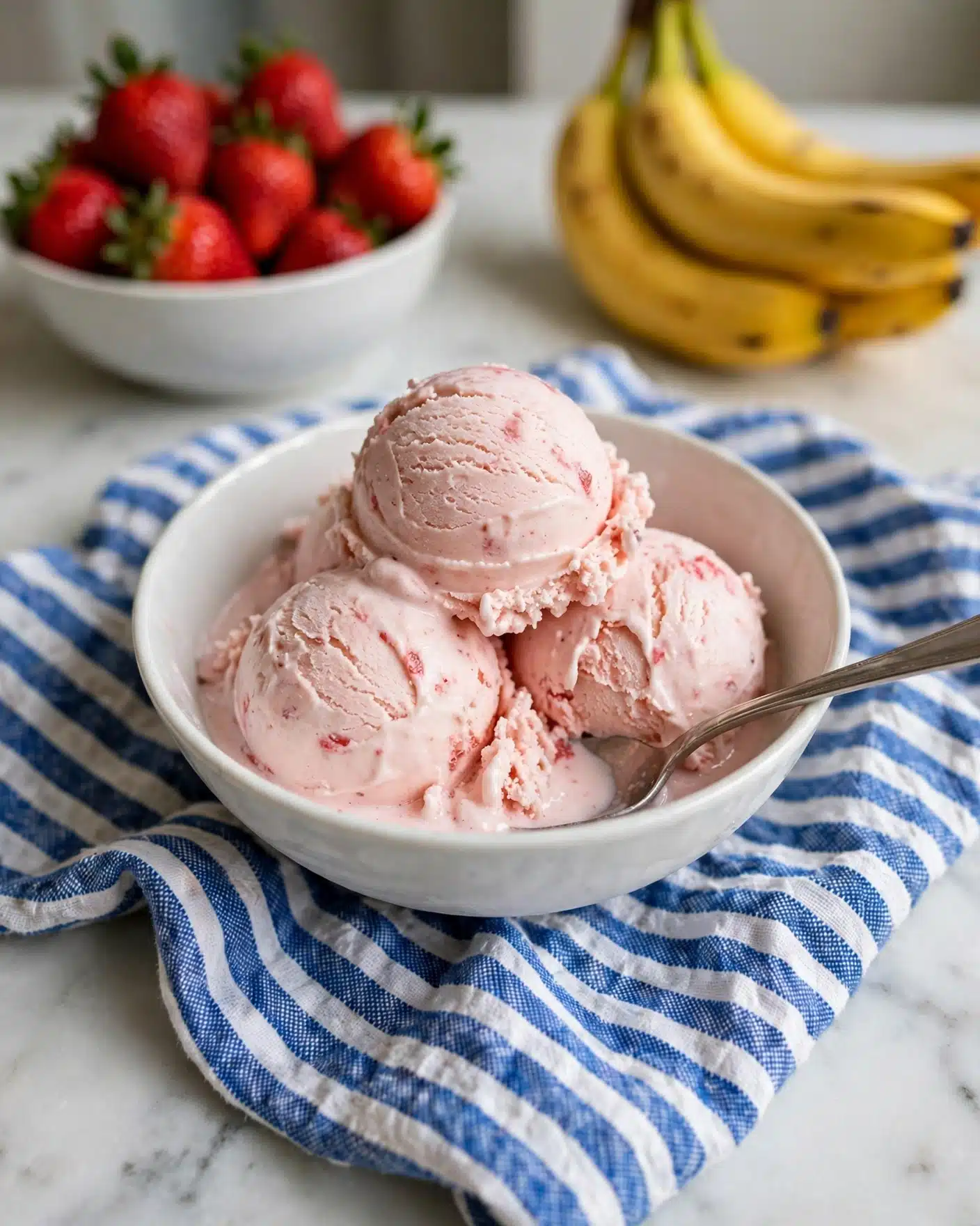 A white bowl is filled with three scoops of light pink strawberry ice cream with a creamy, slightly textured surface. A silver spoon rests inside the bowl, partially submerged in the ice cream. The bowl is placed on a blue striped cloth, all set on a white marbled surface. In the blurred background, there is a white bowl filled with bright red strawberries and a cluster of yellow bananas just visible to the side. Photo taken with an iphone --ar 4:5 --v 7