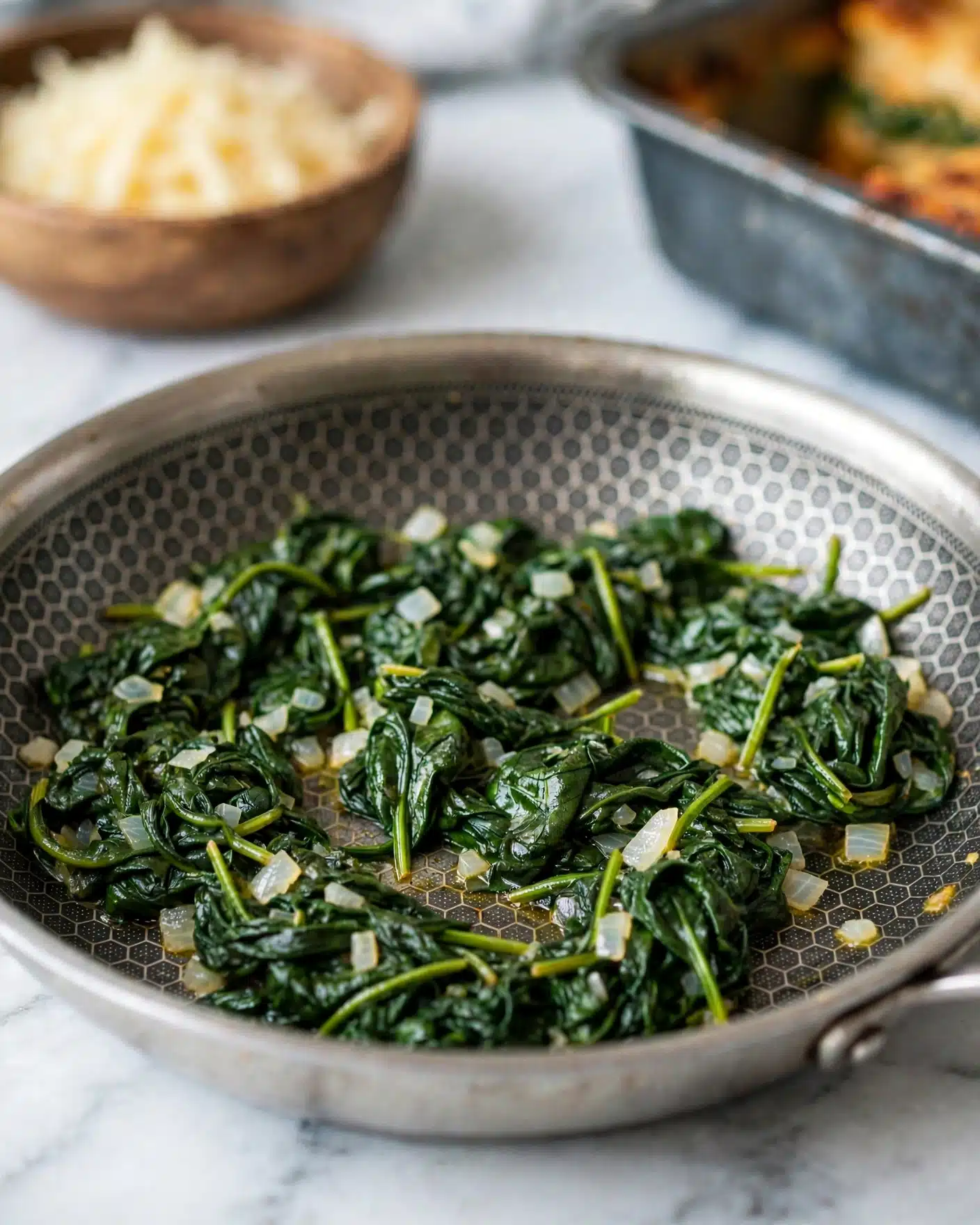 In this image, a silver frying pan with a hexagonal pattern inside holds cooked leafy spinach mixed with small pieces of translucent white onion scattered throughout. The spinach leaves are dark green, soft but still with some texture, and the stems are visible. The pan is on a white marbled surface, and the overall image has a clean and fresh feel. photo taken with an iphone --ar 4:5 --v 7