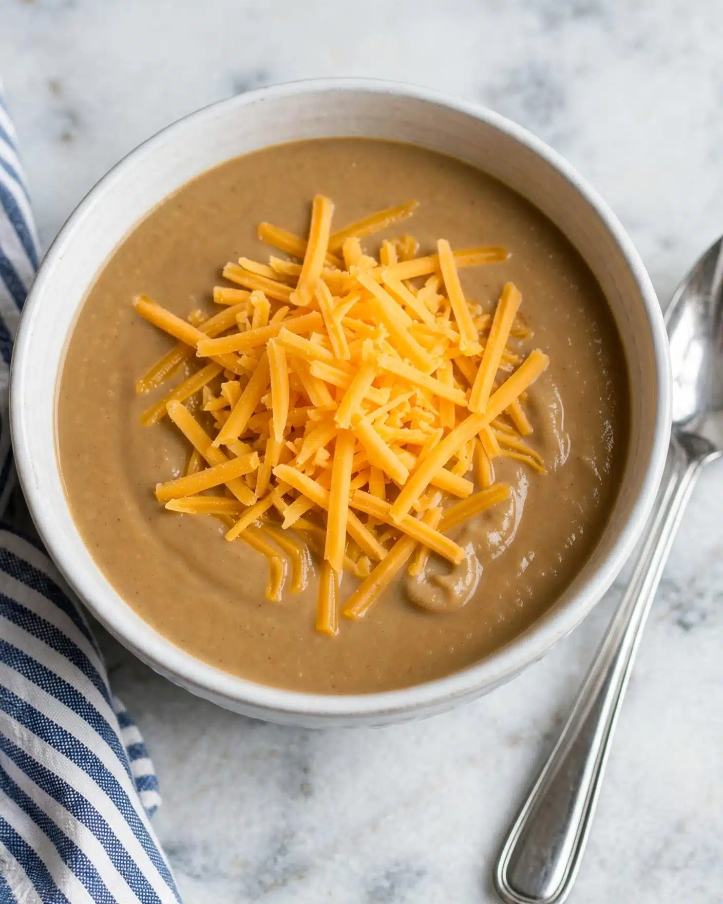 A white bowl filled with a thick, creamy light brown soup as the base layer, topped with a generous pile of shredded bright orange-yellow cheese sitting in the middle. The bowl rests on a white marbled surface next to a long silver spoon and a striped cloth napkin partly visible nearby. The texture of the soup appears smooth and rich, while the cheese shreds add contrast with their sharp edges and vibrant color. photo taken with an iphone --ar 4:5 --v 7
