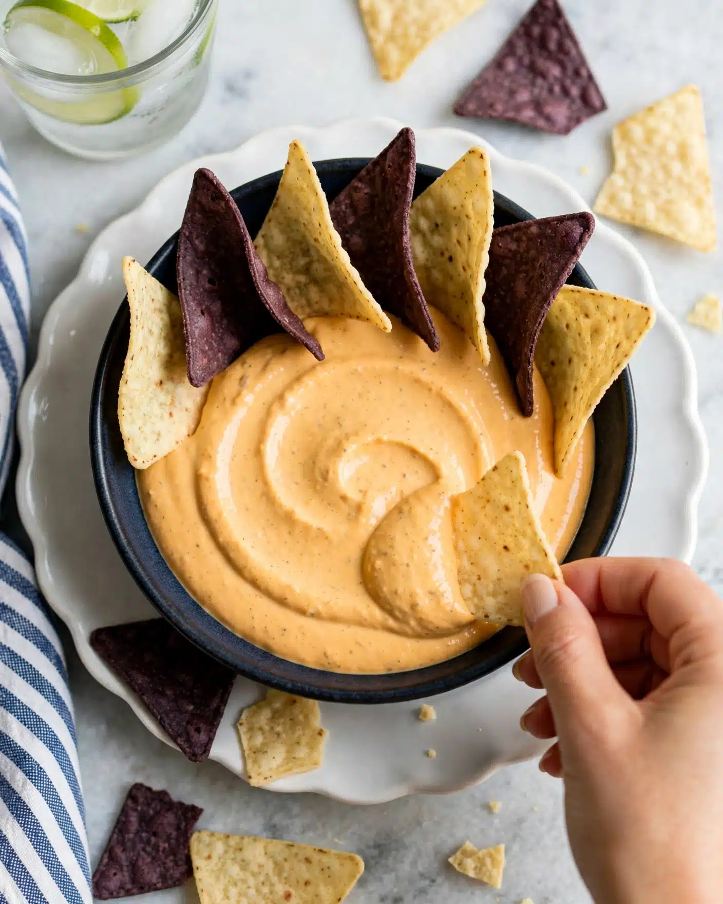 A dark bowl filled with two layers of smooth, light orange cheese dip, topped with a mix of three tortilla chips placed upright around the edge of the bowl; two dark purple chips alternate with three pale yellow chips creating a fan effect. A woman's hand is holding one pale yellow chip, dipping it into the cheese near the bowl’s edge. The bowl sits on a white scalloped plate, which is on a white marbled surface. Scattered around the bowl are additional dark purple and pale yellow tortilla chips, with a clear glass of lime water partially visible on the left side near the top photo taken with an iphone --ar 4:5 --v 7