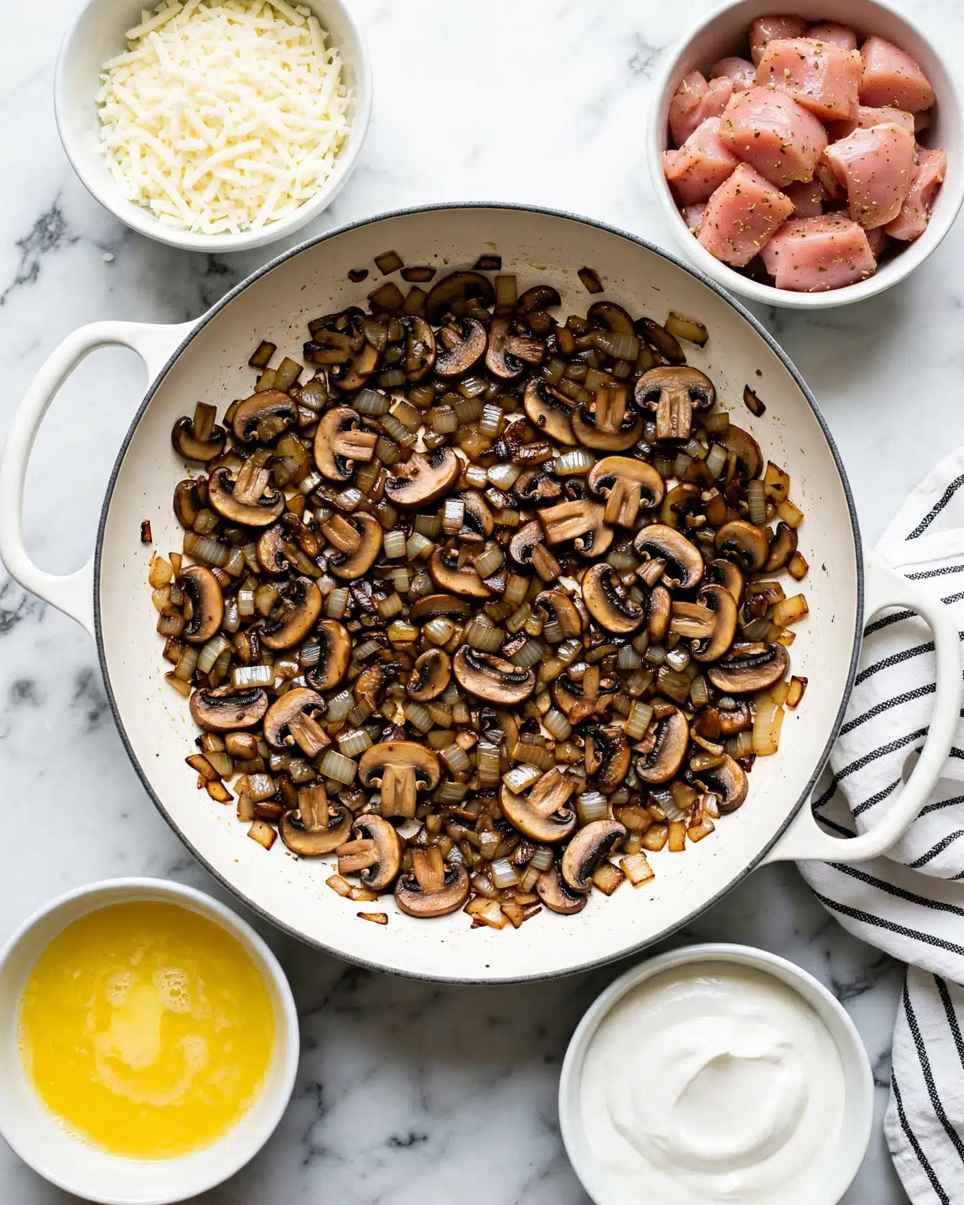 The image shows a white pan with cooked sliced mushrooms and small pieces of browned onions, creating a dark brown and golden mix that covers the base evenly. Surrounding the pan are four white bowls: one filled with shredded mozzarella cheese, another with raw seasoned pink chicken pieces, a third with creamy white sour cream, and the last one with bright yellow melted butter. The surface beneath everything is white marble, and a white cloth with thin black stripes is partly visible under the right handle of the pan. Photo taken with an iphone --ar 4:5 --v 7