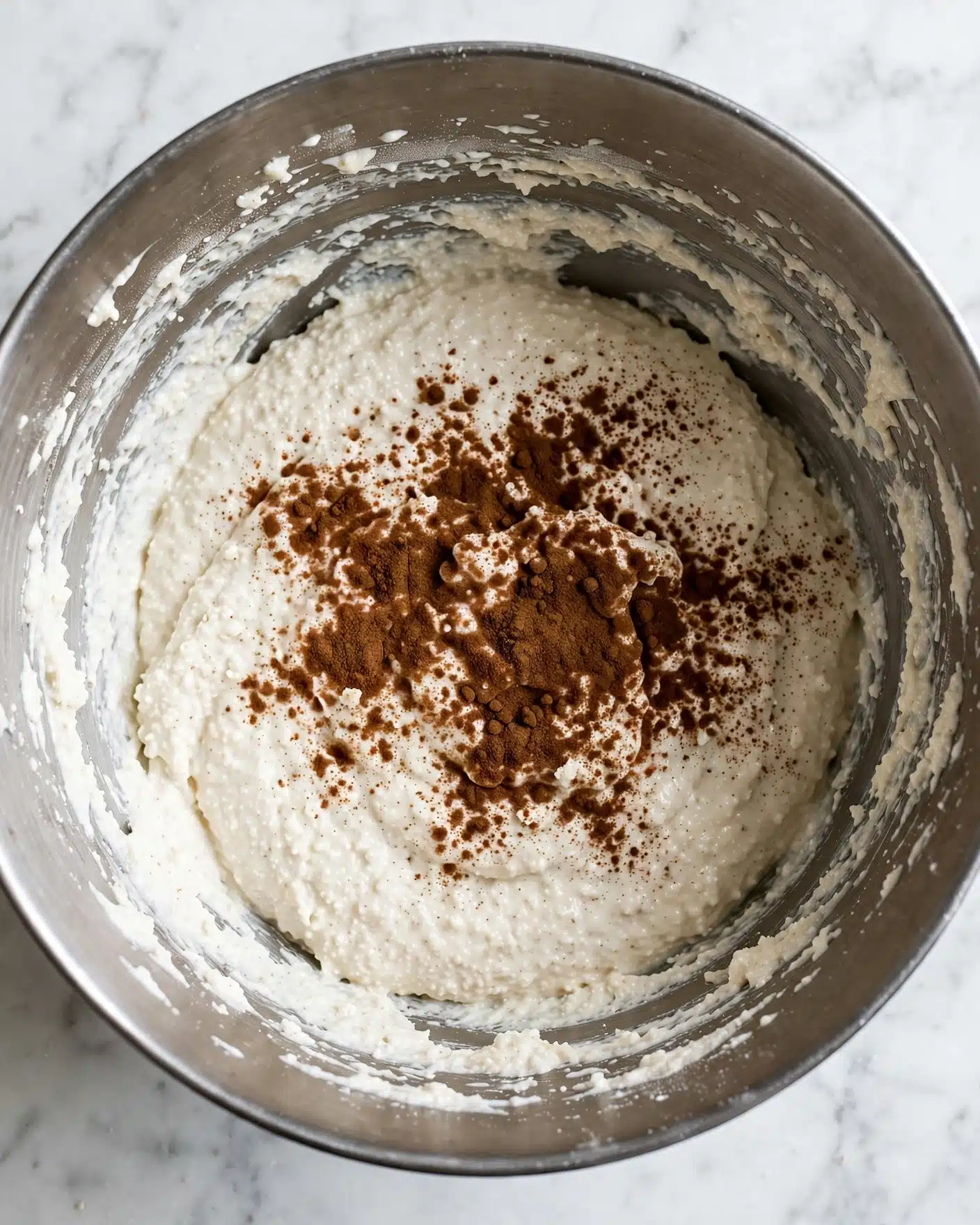 Authentic Cannoli Cream Recipe with Ricotta Recipe 3 A close-up view of a large silver mixing bowl filled with a thick, white creamy batter that has a slightly grainy texture. On top of the batter, there is a generous spread of dark brown cinnamon powder, mostly centered and slightly spread out across the surface. The inside of the bowl shows some batter sticking to the sides, giving it a textured look. The bowl sits on a white marbled surface, and there is soft natural light illuminating the scene, revealing the details of the mixture and spice clearly. photo taken with an iphone --ar 4:5 --v 7