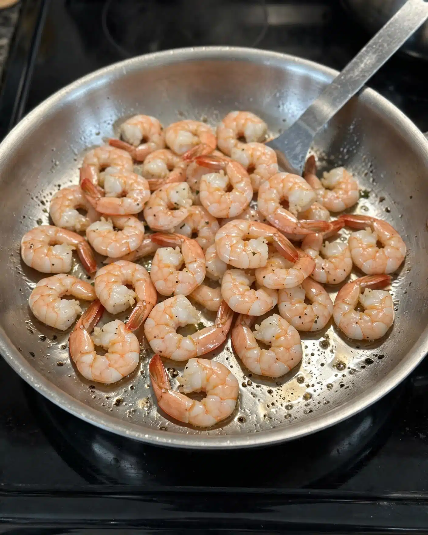 Shrimp Tacos with Spicy Creamy Sauce and Fresh Toppings Recipe 4 A silver pan full of light pink cooked shrimp, some with a few black pepper specks on them, sitting on a black stove. A metal spatula is poking the shrimp from the top right side of the pan. The metal pan surface is shiny with a few small spots of cooking oil and light reflections. photo taken with an iphone --ar 4:5 --v 7