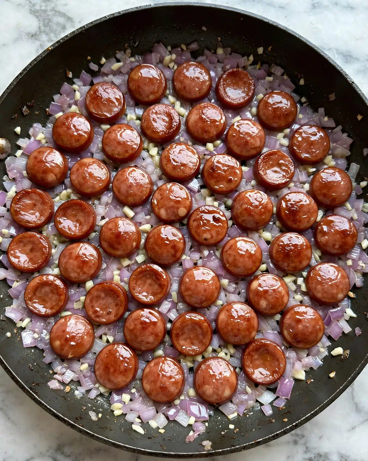 Quick Creamy Chorizo Pasta Recipe 4 The image shows a close-up of a frying pan with two main layers: the bottom layer is small, finely chopped pieces of onion that are light purple and slightly translucent, mixed with tiny bits of white garlic, while the top layer consists of round slices of reddish-brown sausage that have a shiny, cooked surface, all evenly spread across the pan. The background is a white marbled texture visible at the edges of the pan. photo taken with an iphone --ar 4:5 --v 7