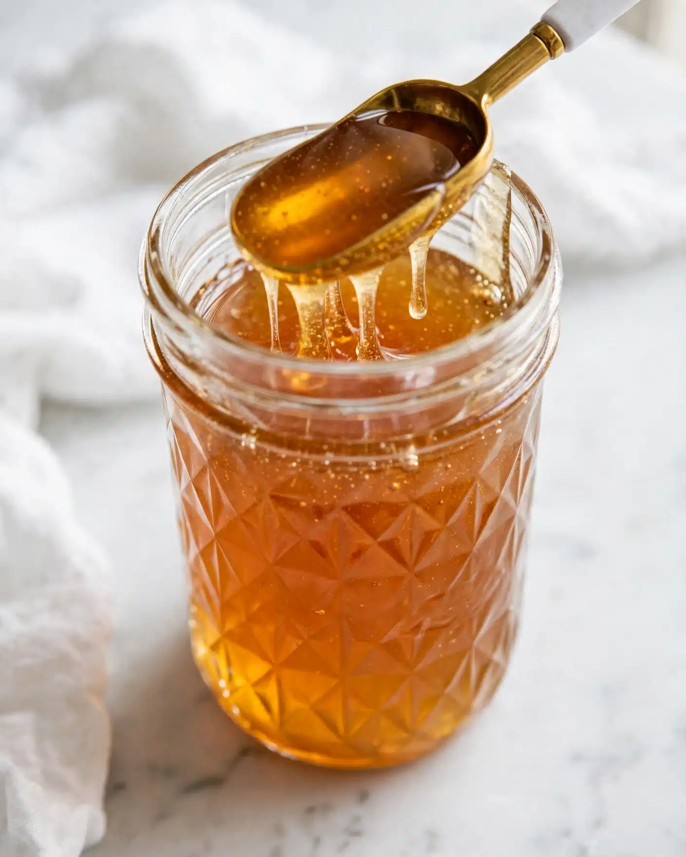 Homemade Caramel Simple Syrup Recipe 5 A close-up of a clear glass jar with a diamond pattern, filled with golden honey. Inside the jar, a spoon with a white handle and gold scoop is dipped in the honey, resting near the top. The jar is placed on a white marbled surface with a soft white cloth partially visible in the background. The honey has a smooth, shiny texture and a warm, amber color. photo taken with an iphone --ar 4:5 --v 7