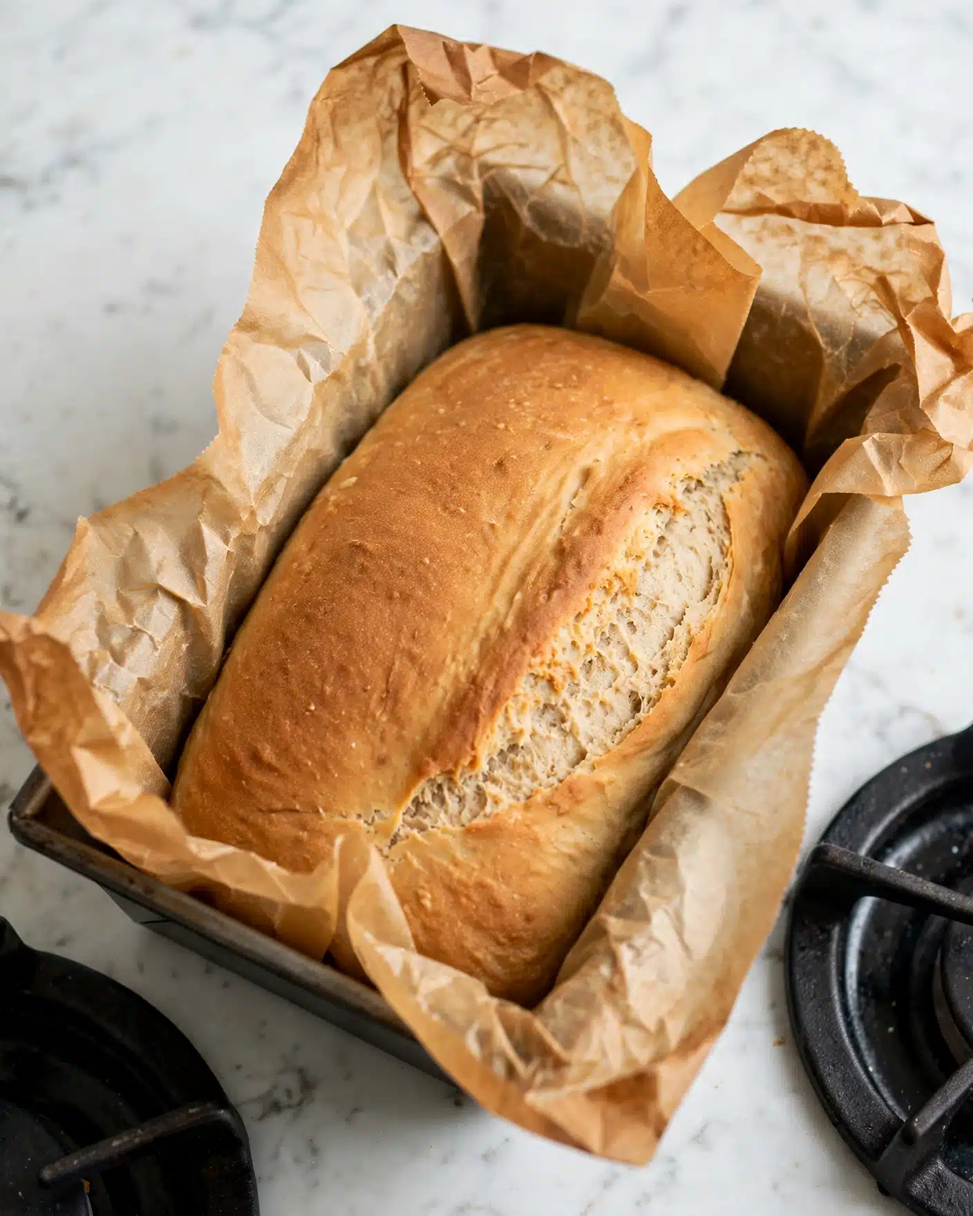 Almond Flour Bread Recipe 5 A single loaf of bread with a smooth, golden-brown top surface sits inside a baking pan lined with crumpled brown parchment paper, which rises above the edges of the loaf. The bread has a slightly cracked texture along one side near the center, revealing a soft, light beige interior. The baking pan rests on a white marbled surface with parts of a black stove burner visible around it. photo taken with an iphone --ar 4:5 --v 7