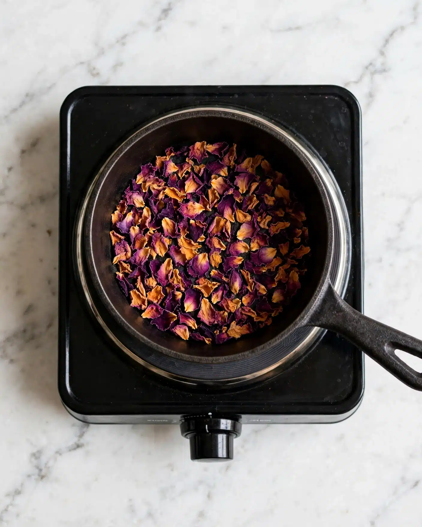 Rose Milk Tea | Rose Tea Latte Recipe 4 A black small pot with a black handle sits on a black electric burner placed on a white marbled surface. Inside the pot, there is a layer of dry rose petals that are mainly dark purple with some orange and yellow edges, spread evenly across the bottom of the pot. The photo is taken from above, showing the pot and burner clearly. photo taken with an iphone --ar 4:5 --v 7