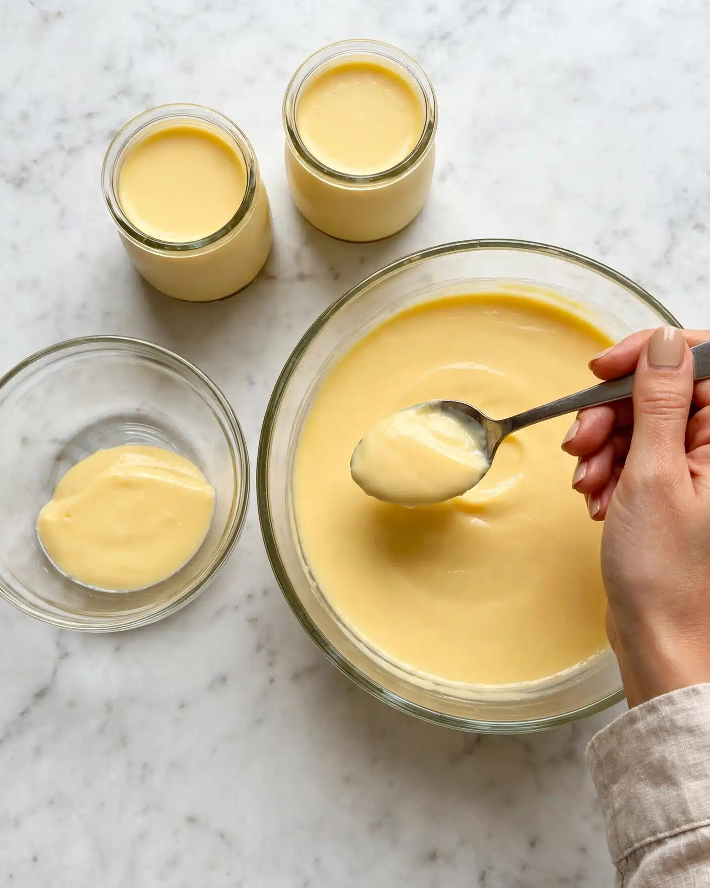 Mango Mousse Recipe 4 The image shows a large clear glass bowl filled with smooth, creamy yellow pudding on the right side, with a woman's hand holding a spoon scooping some pudding from it. Next to it on the left side is a smaller clear glass bowl with a small amount of pudding inside. Above both bowls are two small clear glass jars, filled to the top with the same yellow pudding. The background is a white marbled texture. Photo taken with an iphone --ar 4:5 --v 7