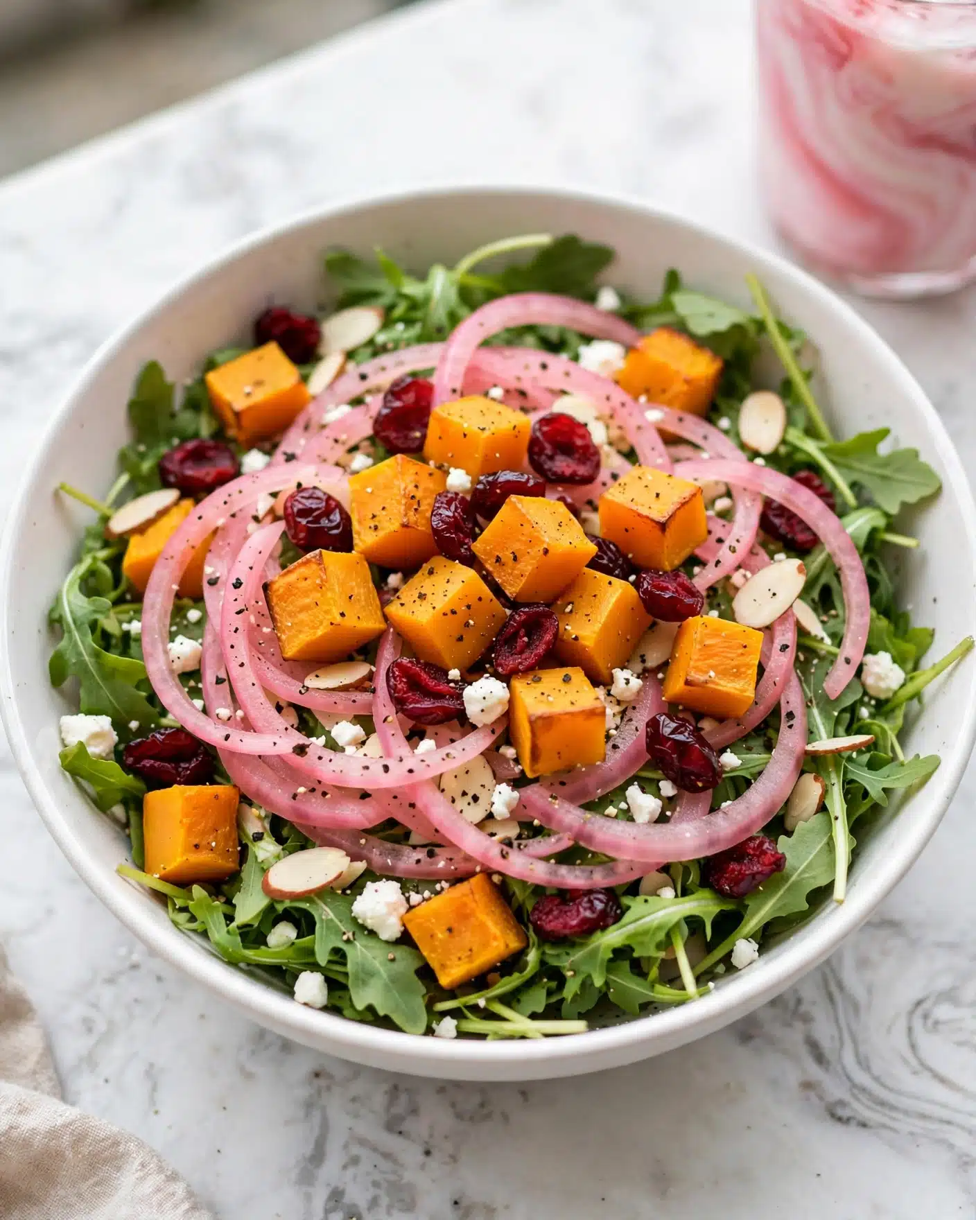 A white bowl filled with a layered salad on a white marbled surface. The base layer is fresh green arugula leaves spread evenly. On top, bright orange roasted butternut squash cubes are scattered along with deep red dried cranberries. Thin, translucent slices of pink pickled red onions are placed across the salad, adding a swirl effect. Small white crumbles of feta cheese and slivered almonds are sprinkled throughout, with a light dusting of black pepper on all ingredients for contrast. In the background, a pink and white marbled drink is partially visible. photo taken with an iphone --ar 4:5 --v 7