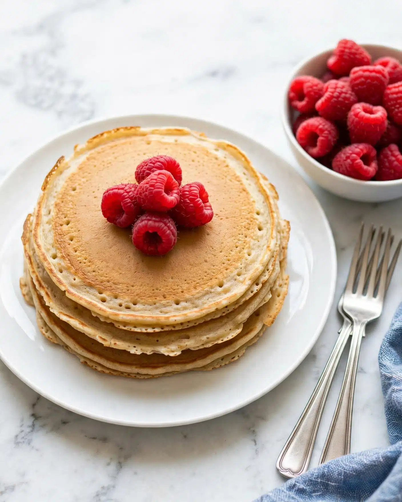 Keto Cream Cheese Pancakes with Almond Flour Recipe 5 A stack of three thin, golden-brown pancakes sits in the center of a white plate, each pancake showing slightly uneven edges and a light, airy texture. On top of the pancakes, there are five bright red raspberries arranged in a small pile. To the right side of the plate, there is a white bowl filled with more raspberries. The setting is on a white marbled surface with two silver forks resting near the plate, and a subtle blue woven cloth partially seen at the edge. Photo taken with an iphone --ar 4:5 --v 7