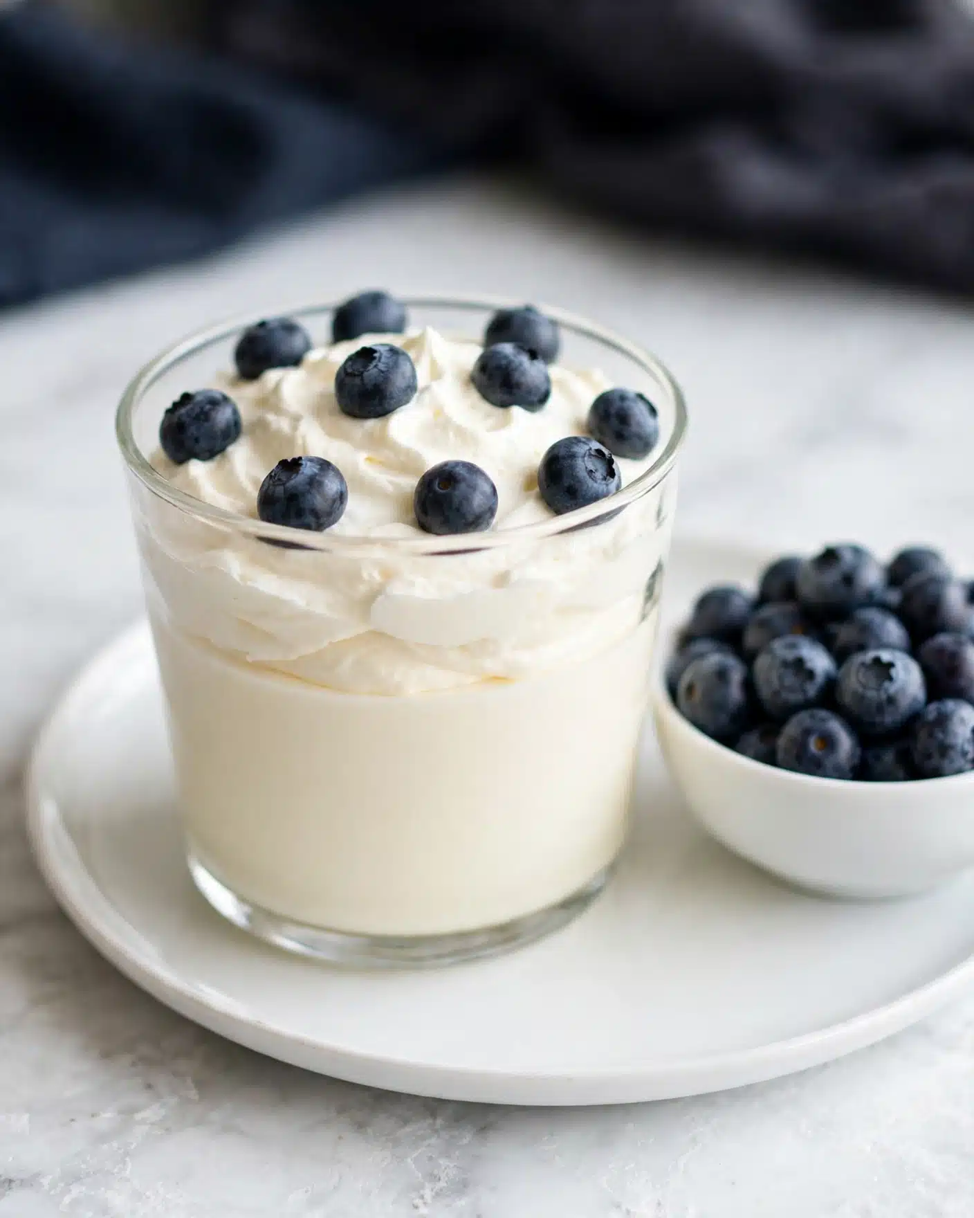 Keto Cheesecake Fluff Recipe 5 The image shows a clear glass filled with two layers: a smooth, creamy white layer at the bottom and a fluffy, swirled white cream layer on top. On the top layer, there are several fresh, round blueberries, dark blue with a slight matte texture scattered evenly. The glass sits on a white plate, and beside it, there is a white bowl filled with more blueberries, both placed on a white marbled textured surface. The background is softly blurred, showing a dark cloth. photo taken with an iphone --ar 4:5 --v 7