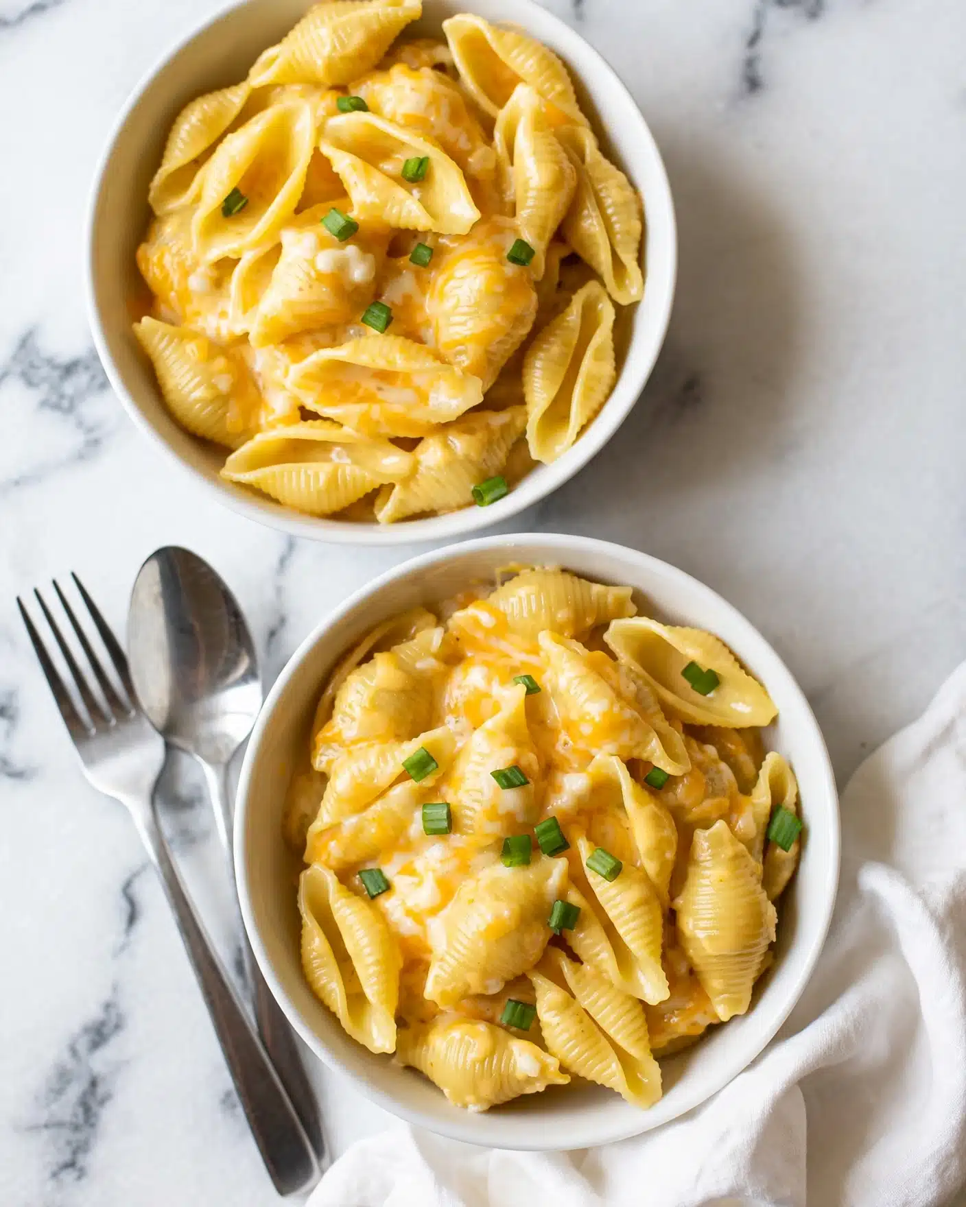Two white bowls filled with creamy yellow pasta shells coated in cheese sauce are placed on a white marbled surface. The pasta has a soft, smooth texture with bits of melted white and orange cheese mixed throughout. Small green onion pieces are scattered on top of the pasta shells in both bowls, adding a touch of color. A silver fork and spoon rest beside the bottom bowl on the left side. A piece of folded white cloth is partly visible at the bottom right corner near the bowls. Photo taken with an iphone --ar 4:5 --v 7