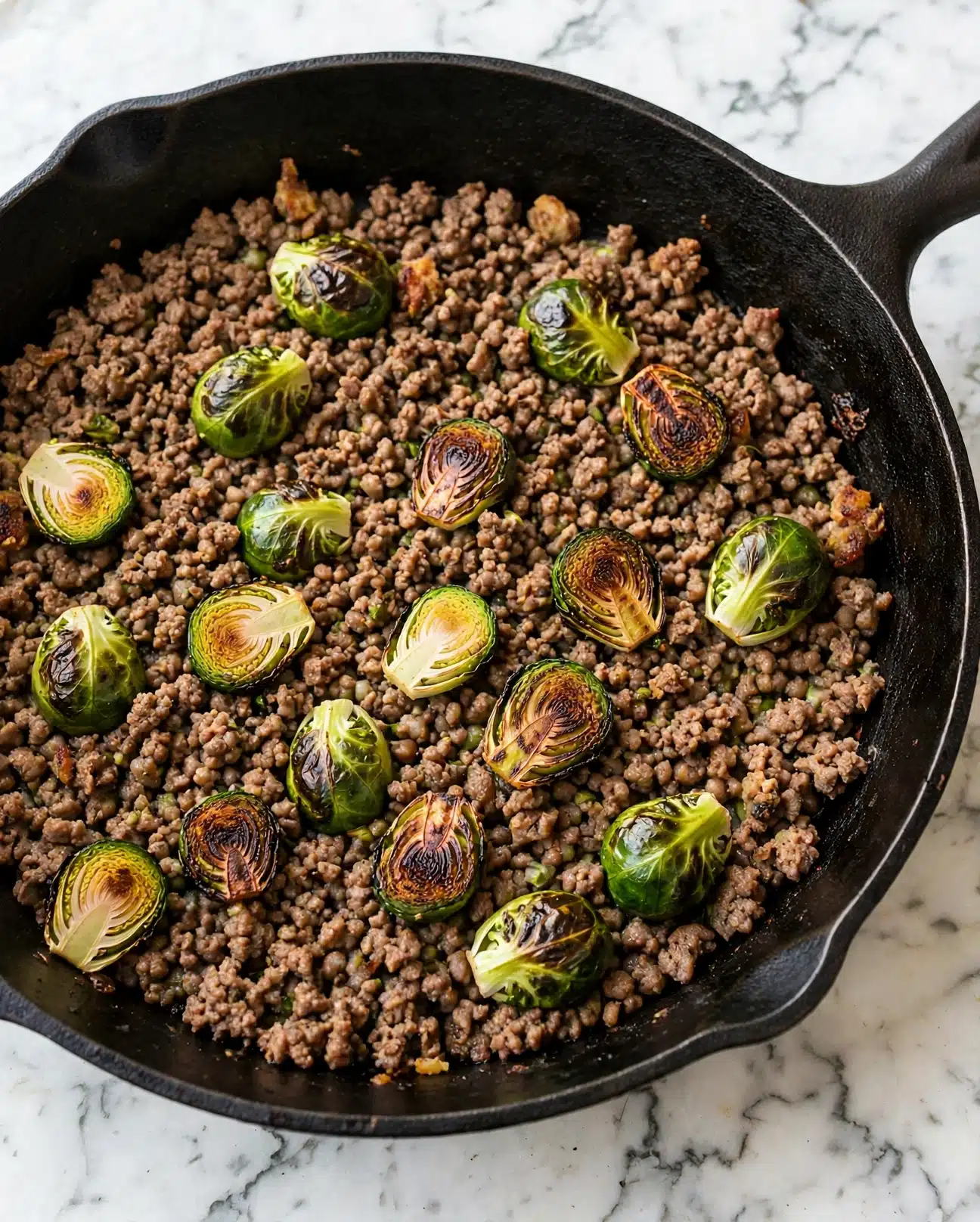 Sweet Chili Ground Beef and Brussels Sprouts Skillet Recipe 5 A close-up view of a black skillet filled with cooked ground meat and halved Brussels sprouts. The meat is brown with some darker, crispy spots, mixed evenly throughout the skillet. The Brussels sprouts show a bright green color with some charred, blackened edges. The skillet sits on a white marbled surface, showing texture slightly visible around the edges. photo taken with an iphone --ar 4:5 --v 7