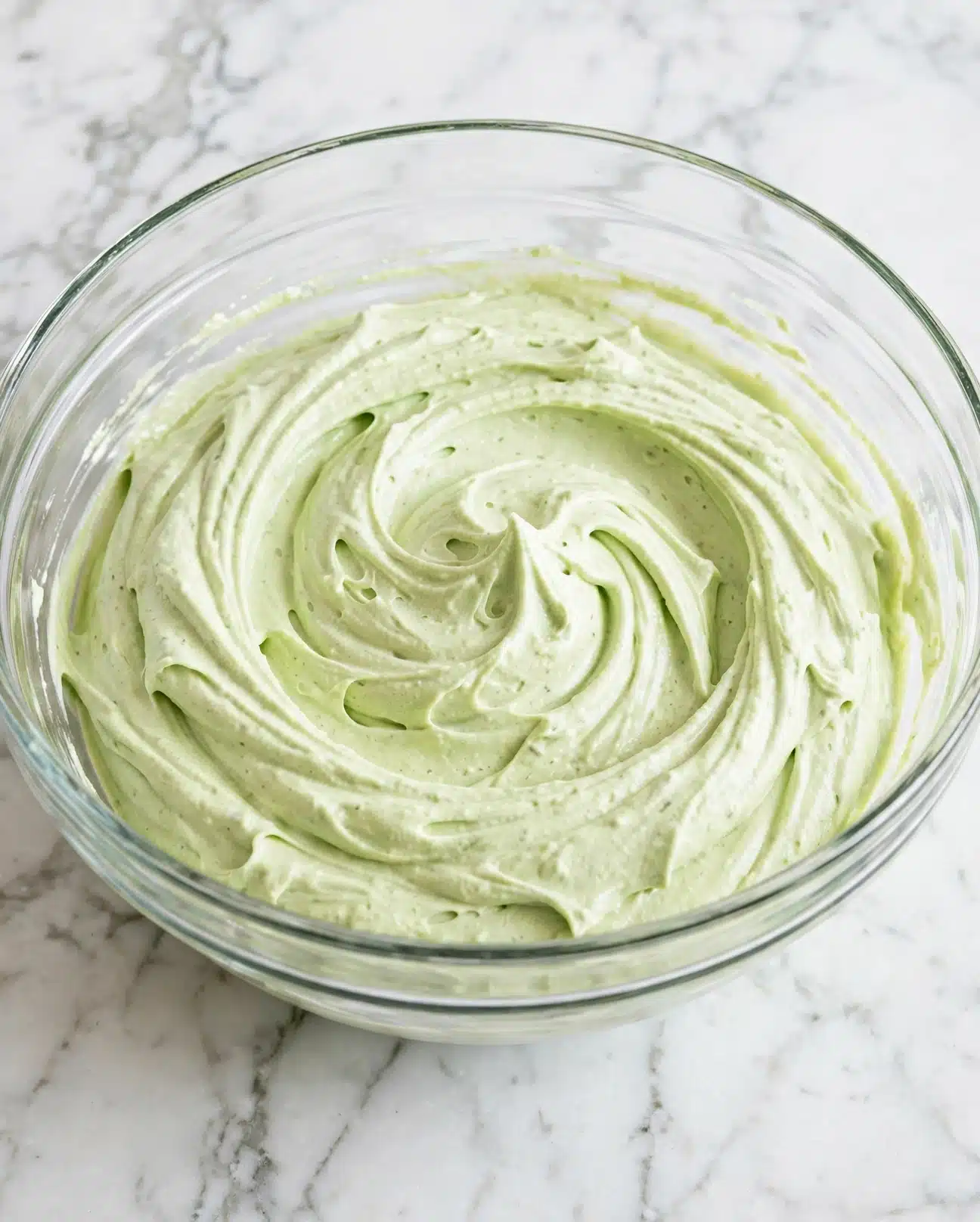 A clear glass bowl filled with a thick, smooth, light green whipped mixture with visible soft swirls and peaks created by stirring. The bowl sits on a white marbled surface. The texture looks creamy and airy, showing detailed ridges from the movement of a whisk or spoon. photo taken with an iphone --ar 4:5 --v 7