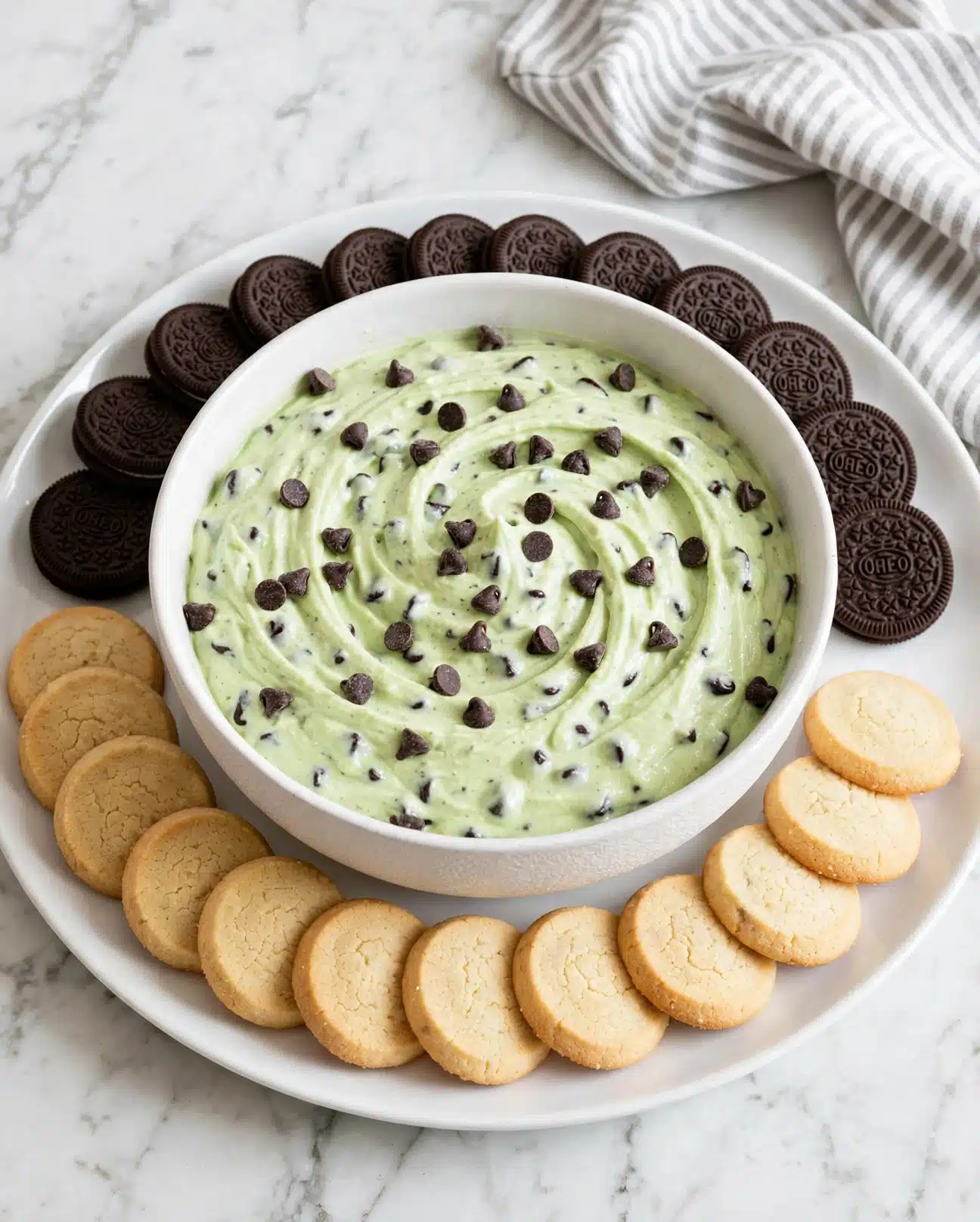A white bowl filled with a light green creamy mixture with small dark chocolate pieces scattered inside and on top. The bowl is placed at the center of a large white plate. Around the bowl, there is a ring of small round vanilla cookies in a light orange color on the bottom half and a ring of dark chocolate sandwich cookies arranged neatly on the top half. The background is a smooth white marbled surface with a folded white and light gray striped cloth visible on the upper right side. Photo taken with an iphone --ar 4:5 --v 7
