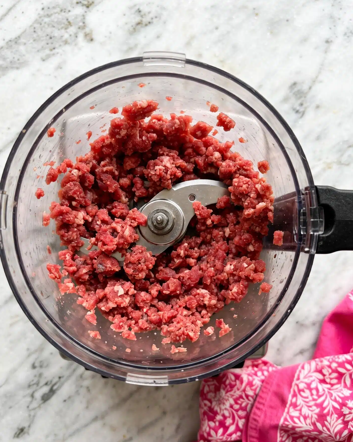No Bake Frosted Animal Cookie Cheesecake Recipe 4 A top view of a food processor bowl filled with minced reddish meat, showing a coarse texture with small uneven chunks all around the central blade. The bowl is transparent with a black handle on the right side. Underneath, there is a white marbled surface and a portion of a bright pink cloth with a white pattern is visible at the bottom right corner. photo taken with an iphone --ar 4:5 --v 7