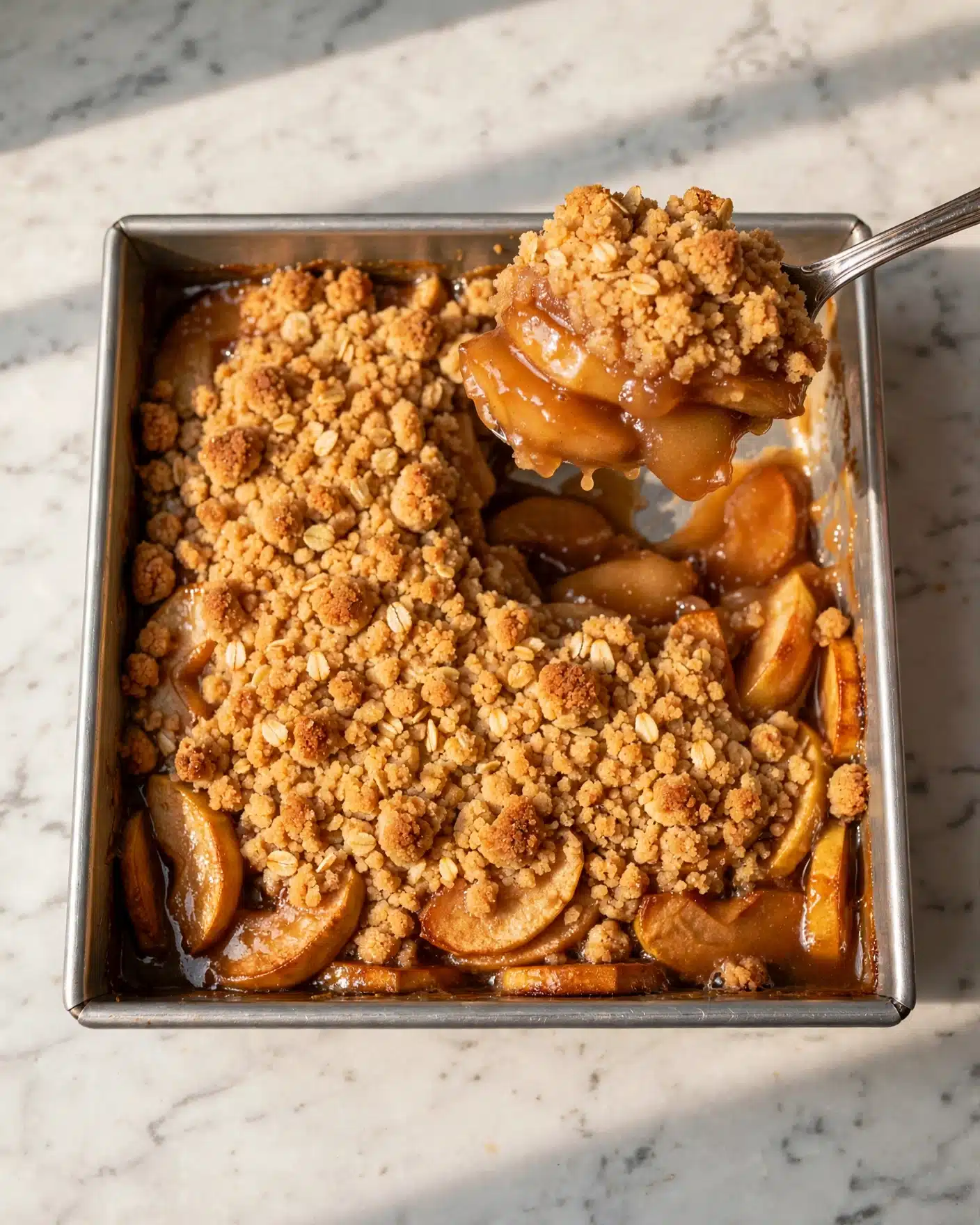 Apple Cream Cheese Dump Cake Recipe 5 A silver spoon holds a thick scoop of dessert showing two layers: the top layer is golden brown with a crumbly, dry texture, and below it is a shiny, soft layer of cooked apple slices in syrup. The dessert sits in a baking dish with a light brown bottom layer of apples and the golden crust layer covering the rest of the dish. The background is a white marbled surface. photo taken with an iphone --ar 4:5 --v 7