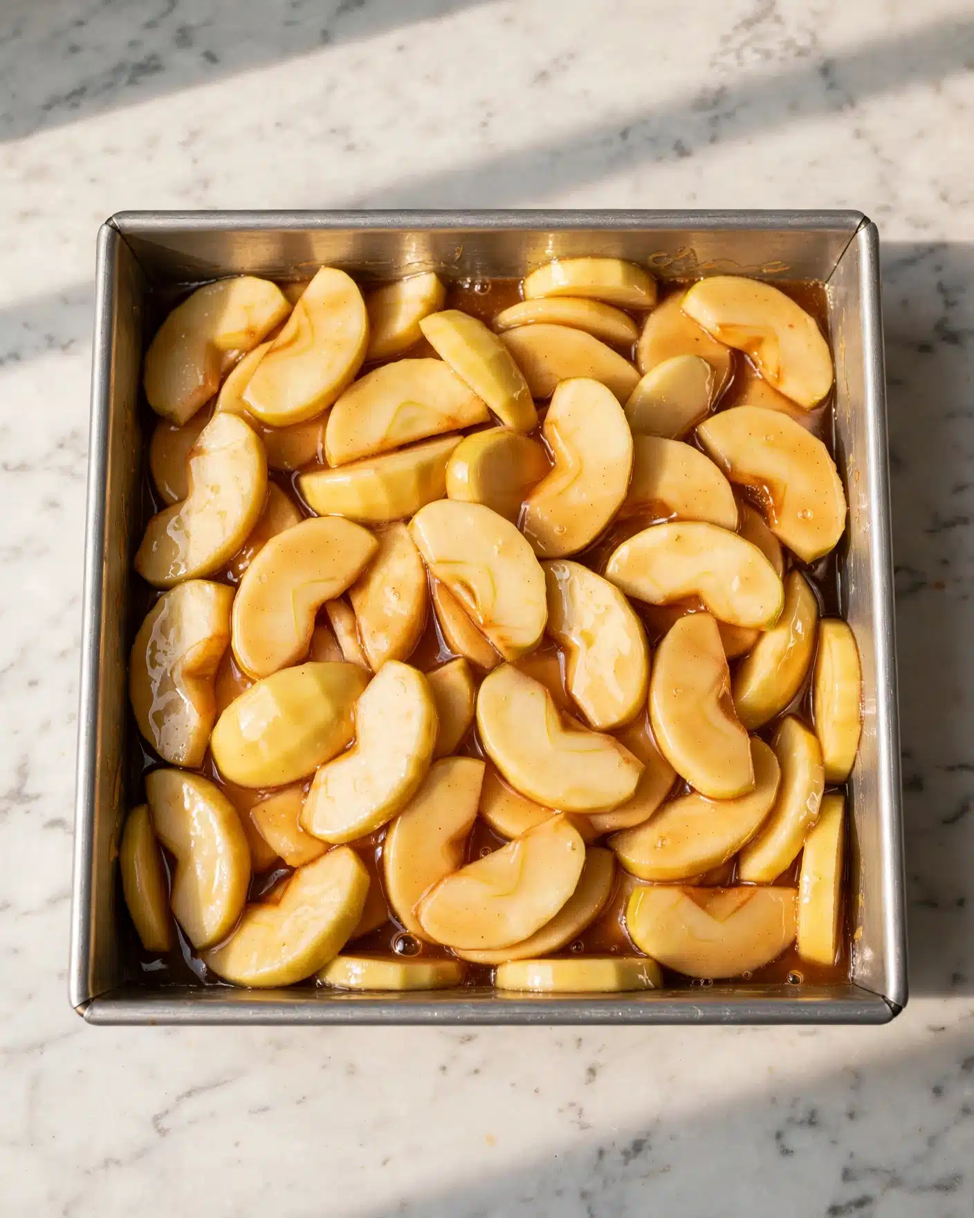 Apple Cream Cheese Dump Cake Recipe 4 A close-up top view of a square silver baking pan filled with a thick mixture of peeled apple slices coated in a golden, shiny syrup. The apple slices vary in size and are layered evenly, filling the pan almost to the top. The pan sits on a white marbled surface that contrasts with the warm colors of the apples and syrup, highlighting the glossy texture of the fruit and syrup blend. photo taken with an iphone --ar 4:5 --v 7