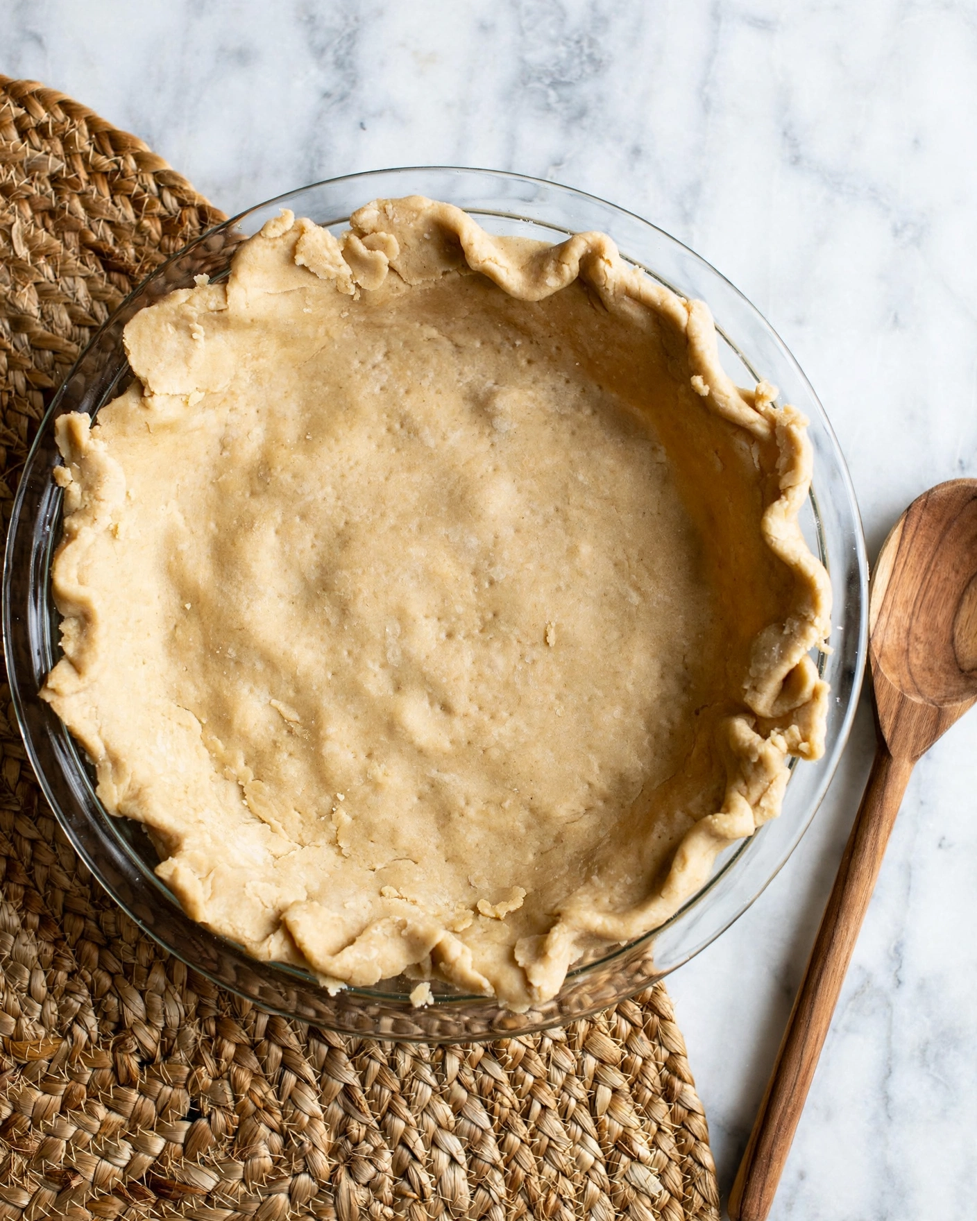 S'mores Pie With Marshmallow Fluff Recipe 4 A round clear glass pie dish shows one thick, even layer of light tan pie crust pressed smoothly against the bottom and sides, with slight uneven edges folded over the rim. The pie sits on a textured woven mat with warm natural tones. To the right is a wooden spoon with a smooth surface and soft wood grain. The photo is taken from above on a white marbled surface. photo taken with an iphone --ar 4:5 --v 7