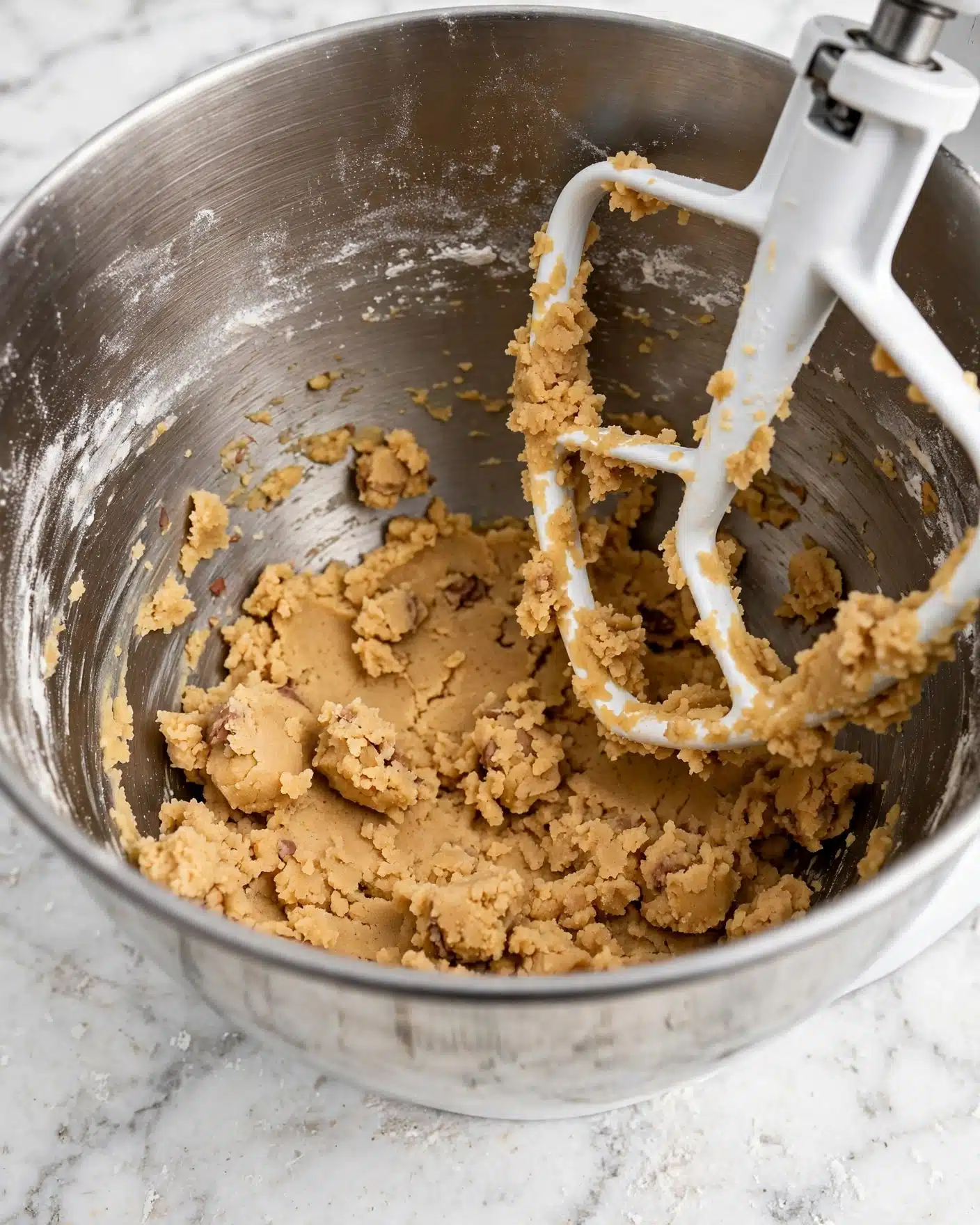 Gingerbread Cookie Bars Recipe 4 A close-up image of light brown cookie dough inside a stainless steel mixing bowl with a white mixer paddle attachment partially covered in the dough. The dough appears soft and slightly chunky, spread unevenly across the bottom and sides of the bowl. The inside surface of the bowl shows some flour residue dusting, adding texture contrast to the smooth metal. The scene is set on a white marbled textured surface. Photo taken with an iphone --ar 4:5 --v 7