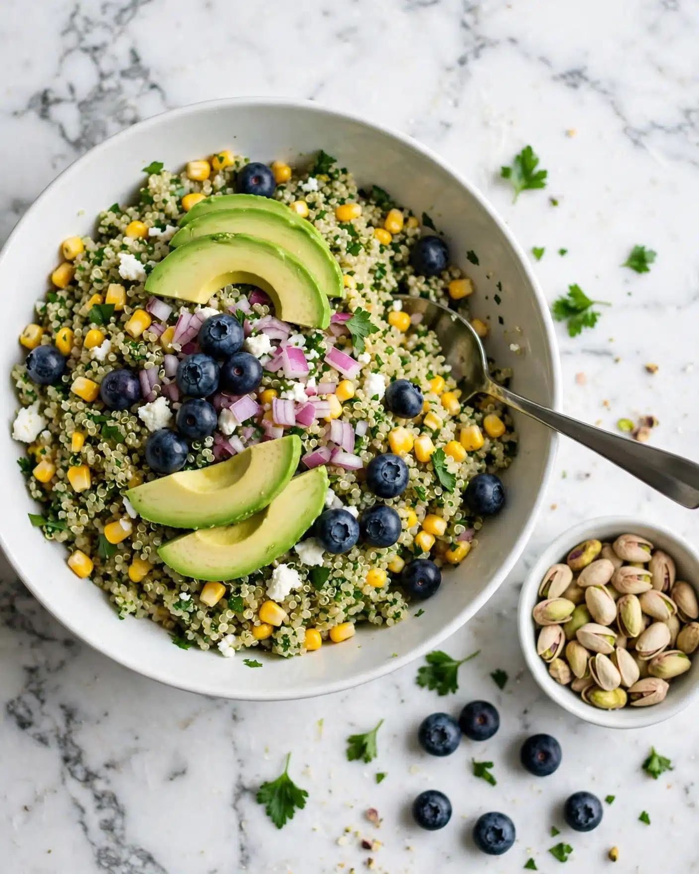 Avocado Blueberry Quinoa Salad with Feta and Cilantro Lime Dressing Recipe 5 A large white bowl filled with a colorful quinoa salad is set on a white marbled surface. The salad has a base layer of light green quinoa mixed with bright yellow corn kernels and small bright green herbs. Scattered on top are fresh dark blue blueberries, small white cheese crumbles, and finely chopped red onions showing purple bits. Slices of green avocado are placed on top in several areas, adding smooth texture and bright color. A metal spoon rests in the bowl, ready to serve. Nearby, a small white bowl holds light green pistachios, and a few blueberries and herb leaves are scattered on the surface. photo taken with an iphone --ar 4:5 --v 7