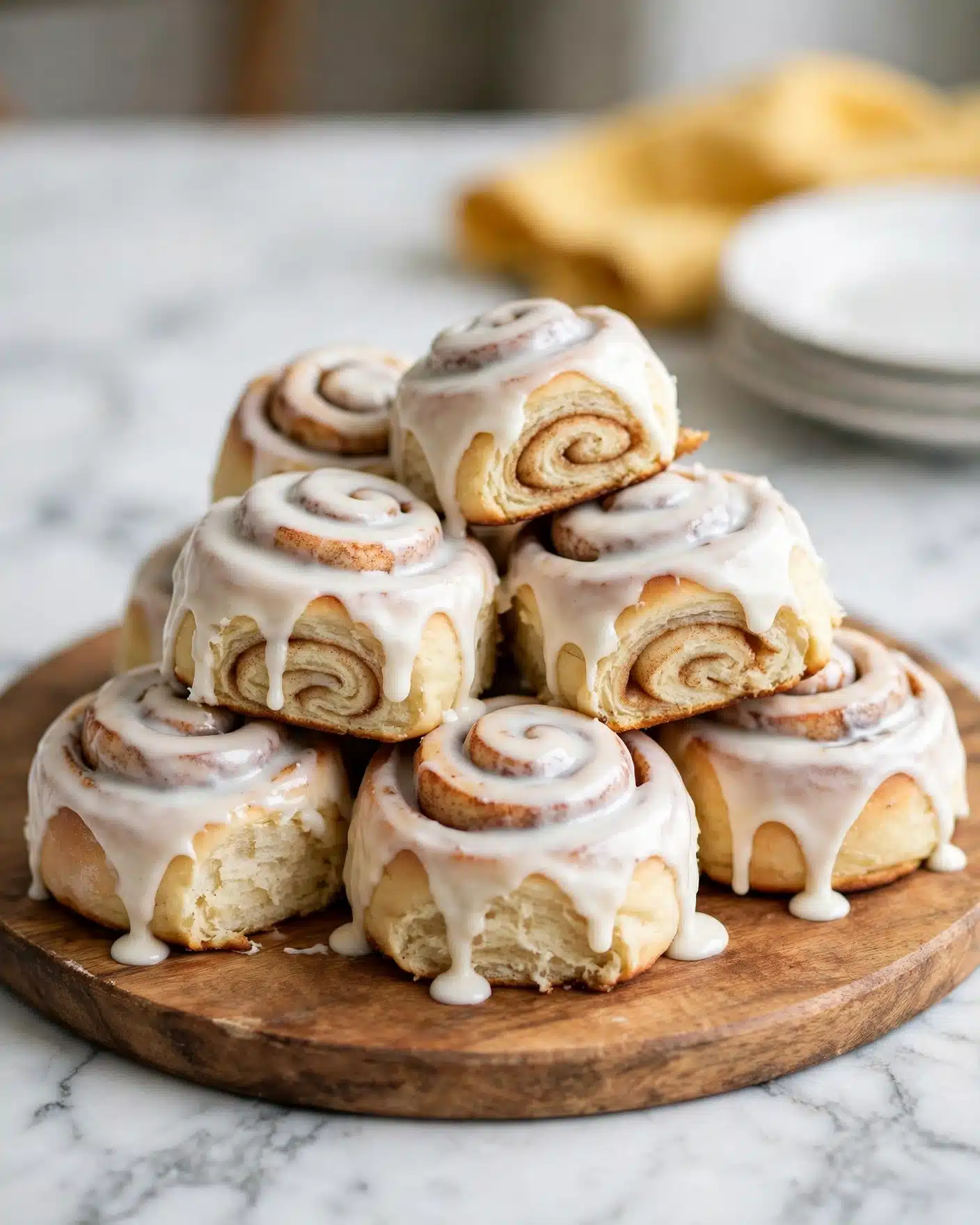Easy Homemade Honey Buns Recipe 5 A small pile of seven cinnamon rolls covered in smooth white icing sits on a round wooden board. Each roll shows soft, swirled layers beneath the icing, with light brown dough peeking through in places. The rolls appear fluffy and thick, arranged loosely in a small pyramid shape. The background features a white marbled surface, with a yellow cloth and some stacked white plates blurred in the distance. Photo taken with an iphone --ar 4:5 --v 7
