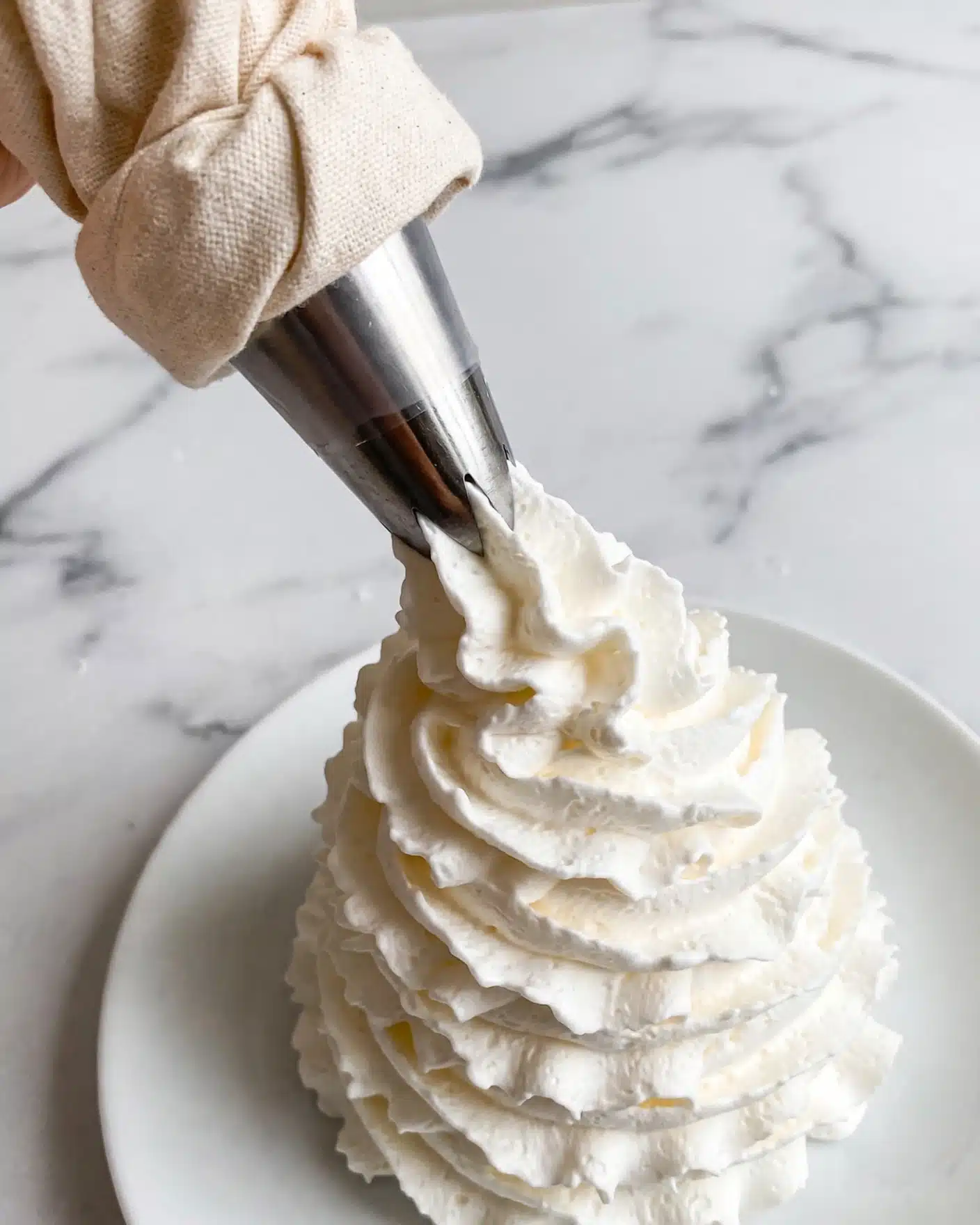 Marshmallow Fluff Recipe 5 A swirl of white whipped cream is being piped onto a smooth white plate using a metal piping bag nozzle. The whipped cream forms one thick, textured mound with visible ridges spiraling up from the base to the tip. The background and surface are a clean white marble. The image focuses closely on the whipped cream and piping bag tip. photo taken with an iphone --ar 4:5 --v 7