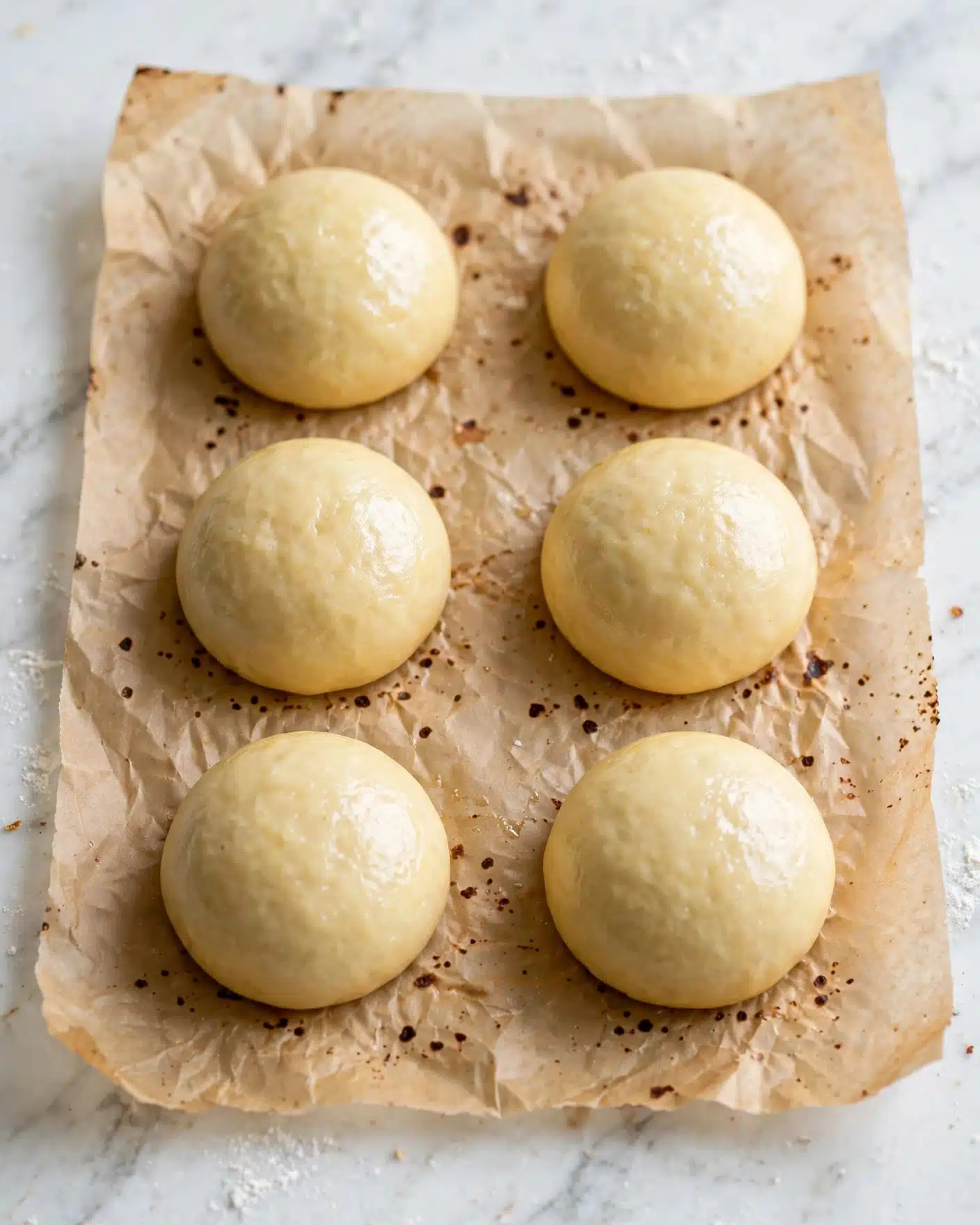 Italian Peach Cookies Recipe 4 The image shows six smooth, round dough balls evenly spaced on a sheet of parchment paper that rests on a white marbled surface. The dough balls are light yellowish in color with a soft look, each having a slightly shiny texture on the top. The parchment paper has some scattered dark spots and creases, while the background remains clean and bright, emphasizing the dough balls' soft, smooth shape and uniform size. Photo taken with an iphone --ar 4:5 --v 7