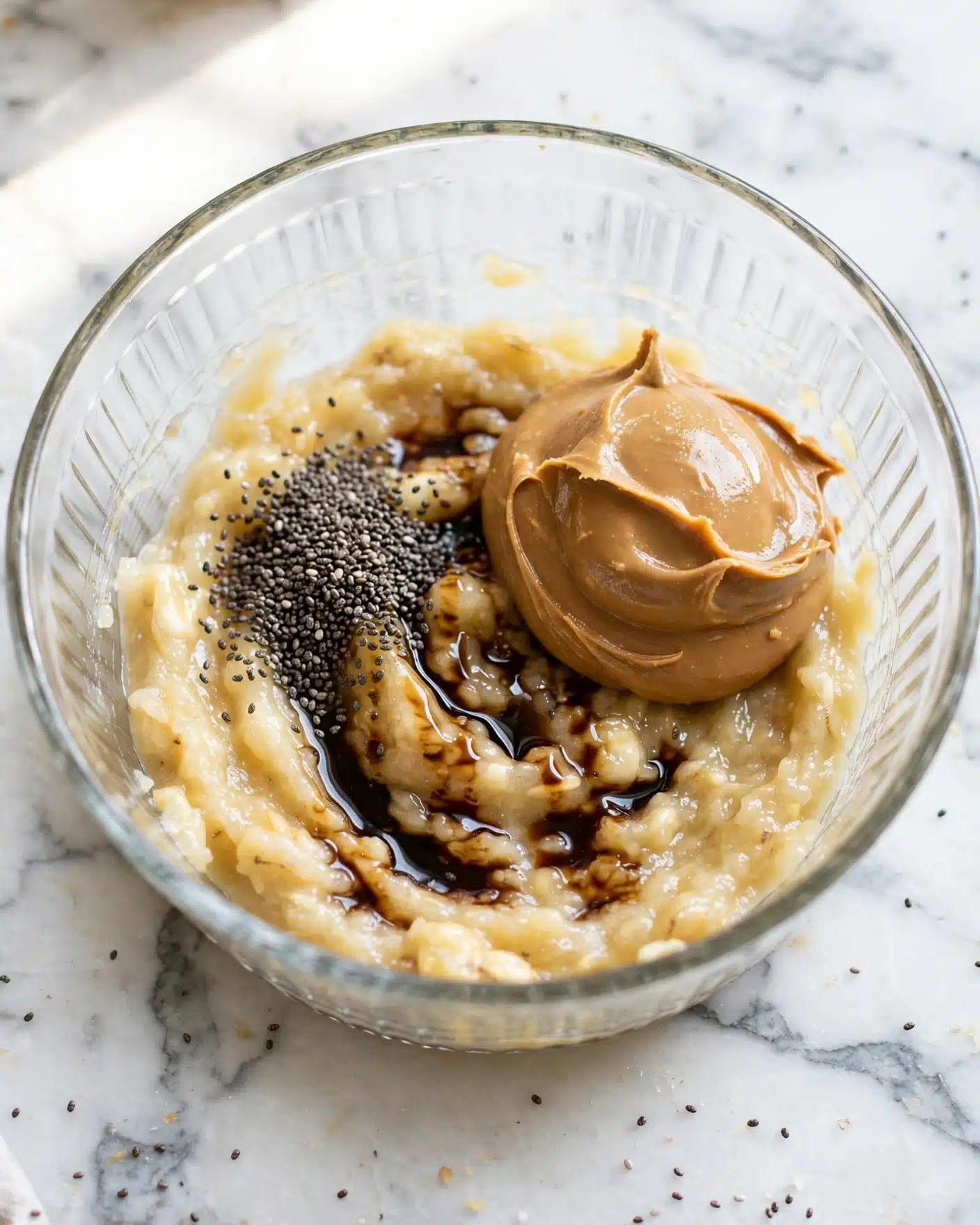 Easy Banana Baked Oatmeal Recipe 4 In the clear glass bowl on a white marbled surface, there is a mix of ingredients grouped closely. At the bottom, there is a creamy mash of light yellow bananas. On top, slightly to the right, there is a smooth, thick scoop of light brown peanut butter. Near the center, there is a layer of dark brown vanilla extract drizzled over the bananas. Next to it, a pile of fine black chia seeds sits loosely on the banana base. The bowl shows clear texture of the mashed bananas and the creamy peanut butter, all lit by natural light. photo taken with an iphone --ar 4:5 --v 7