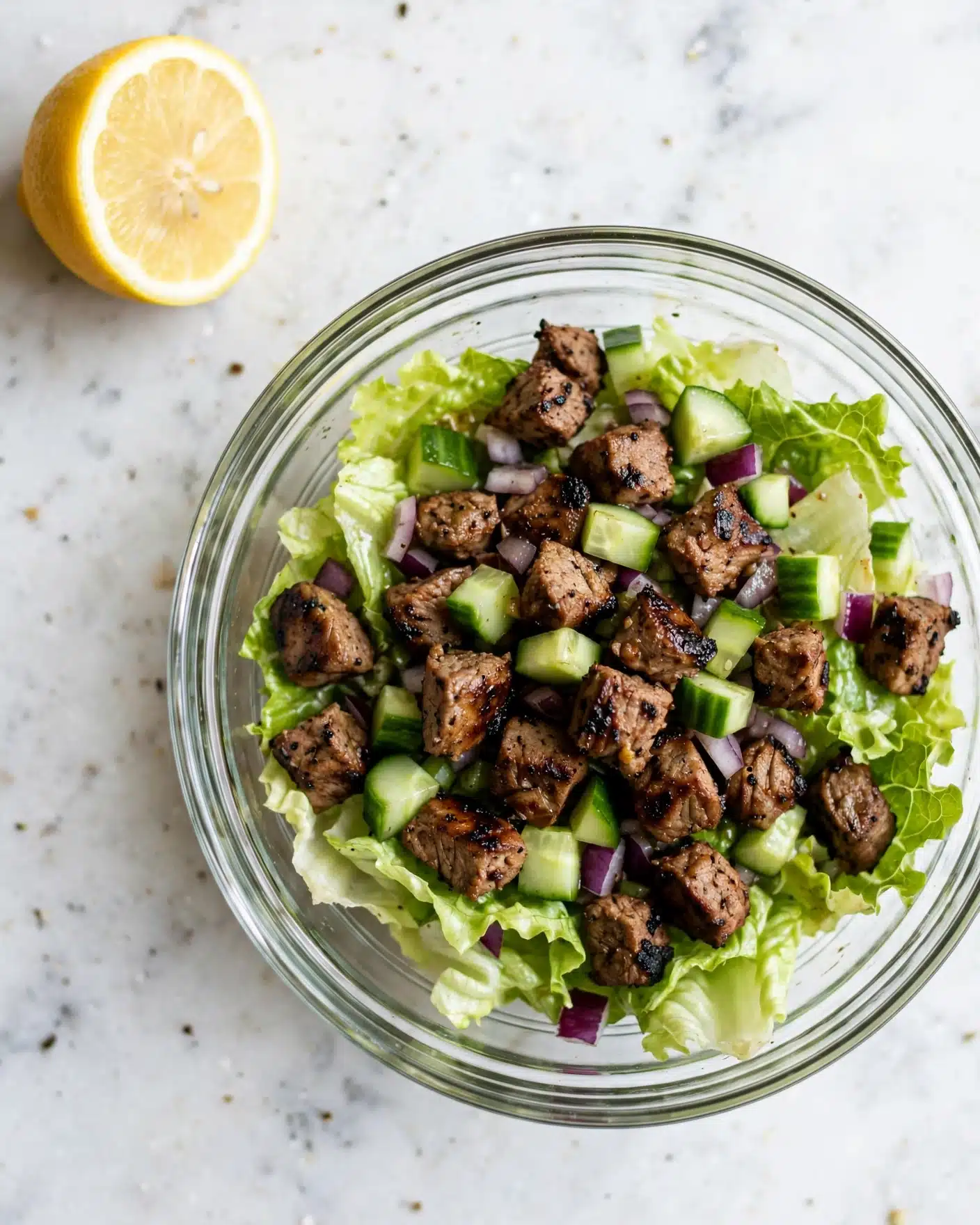 A clear glass bowl sits on a white marbled surface, filled with a fresh green salad. The bottom layer is made of light green, slightly crinkled lettuce leaves. Scattered on top are small bright green cucumber pieces with a smooth texture and medium brown, grilled cubes of meat with dark char marks. There are also small pieces of diced purple-red onion mixed throughout. In the top left corner of the image, a lemon half with a bright yellow rind and pale yellow flesh rests on the white marbled surface. photo taken with an iphone --ar 4:5 --v 7