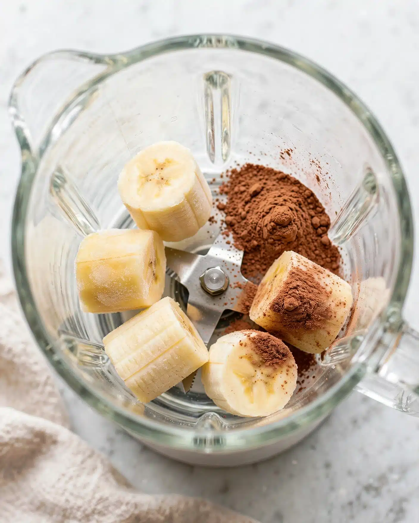 Creamy Banana Cinnamon Smoothie Recipe 4 Inside a clear blender container, there are five pieces of light yellow frozen banana chunks arranged with three on the left side and two on the bottom right. On the top right, there is a small pile of brown cocoa powder, spreading slightly onto the bananas. The container sits on a white marbled surface, with soft natural light illuminating the scene. photo taken with an iphone --ar 4:5 --v 7
