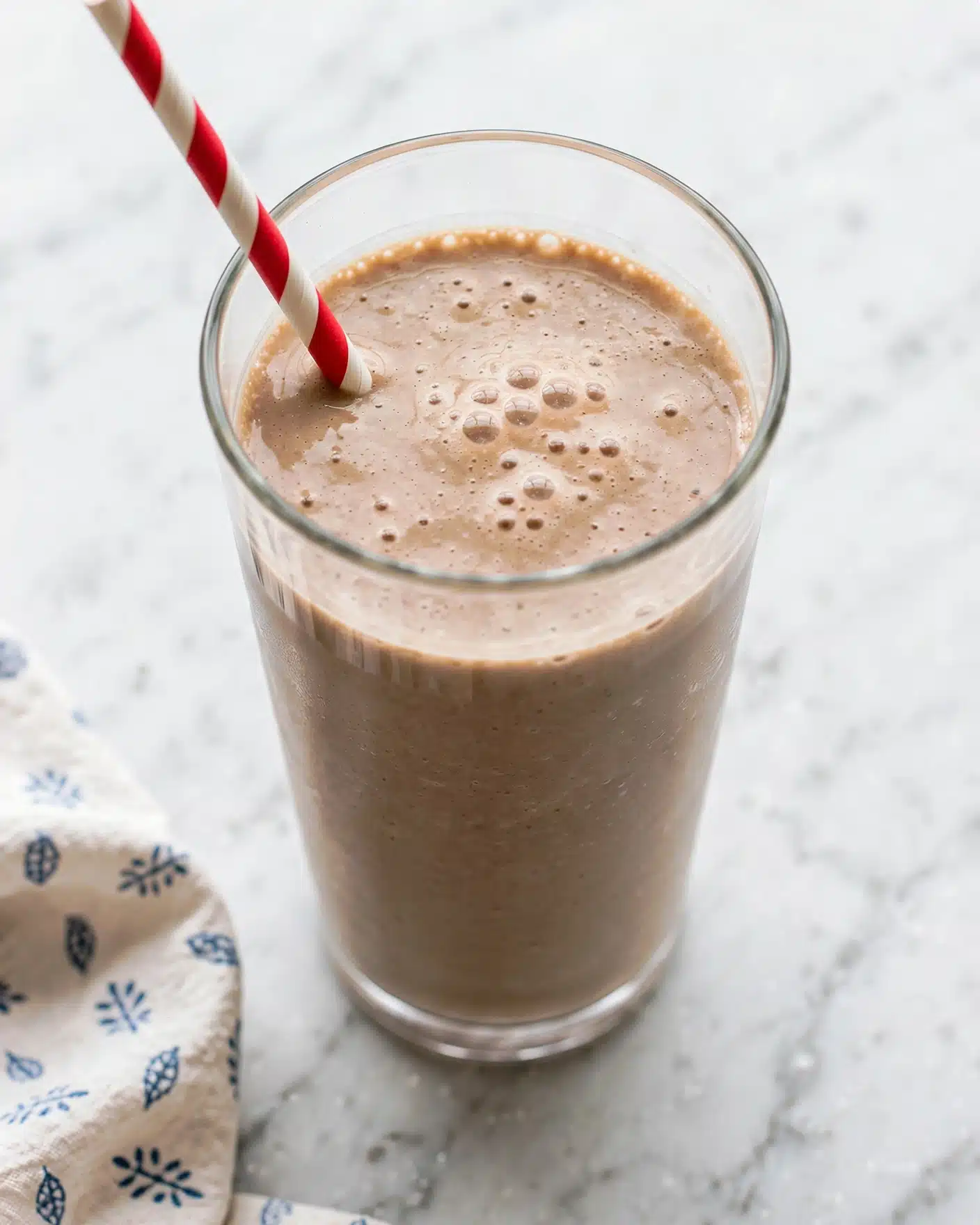 Creamy Banana Cinnamon Smoothie Recipe 5 A tall clear glass filled with a light brown smoothie with small bubbles on the top layer, showing a creamy and slightly frothy texture. A red and white striped paper straw is inserted in the smoothie from the top left side, casting a shadow inside the glass. The glass is placed on a white marbled surface that reflects soft natural light, with a pale cloth featuring a simple blue pattern folded next to it. Photo taken with an iphone --ar 4:5 --v 7