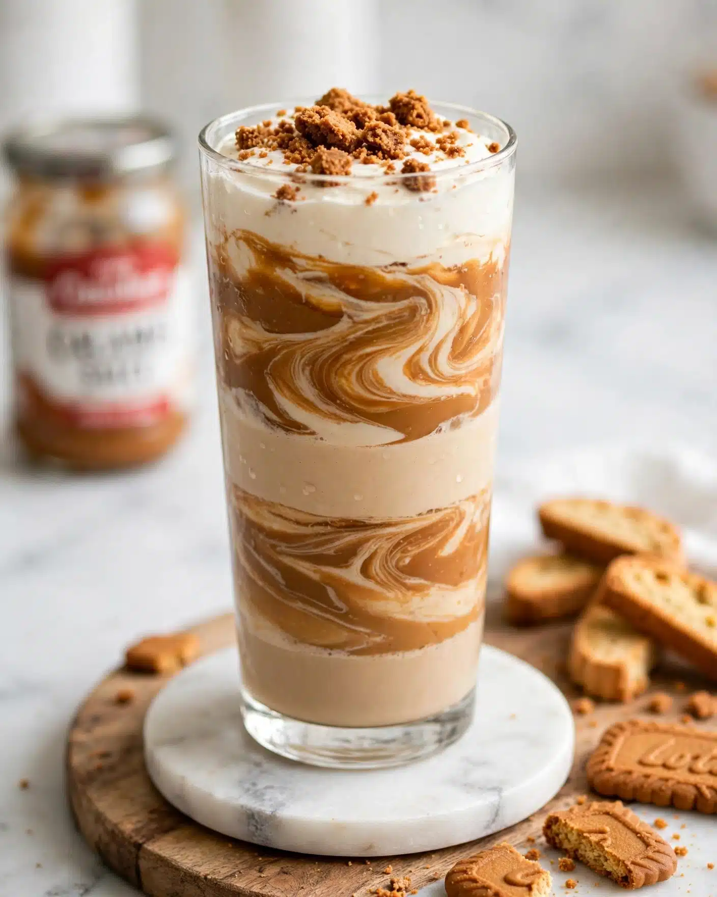Iced Biscoff Latte with Cold Foam Recipe 5 A tall clear glass sits on a round white marble coaster, showing five visible layers of a creamy drink and a thick caramel-colored spread swirling together in a marbled pattern. The bottom layer is a light tan liquid, then a thick caramel layer with some cream peeking through, followed by another light tan creamy layer, then a bold caramel layer mixed with cream, and finally topped with a smooth cream layer sprinkled with crumbled brown biscuit crumbs. The glass is placed on a wooden board with some orange-brown biscuit pieces and a jar with a red and white label blurred in the background, all set on a white marbled surface. photo taken with an iphone --ar 4:5 --v 7