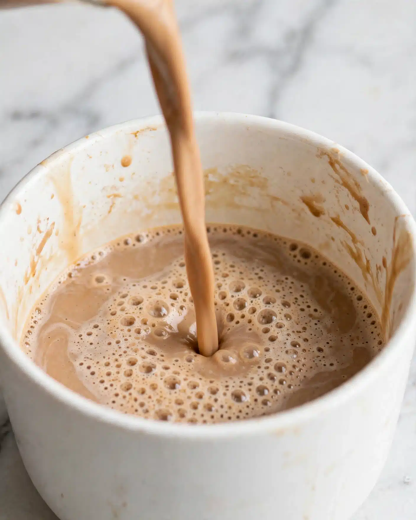 Iced Biscoff Latte with Cold Foam Recipe 4 The image shows a close-up of a creamy brown liquid being poured into a white container. The surface of the liquid has small bubbles and a slightly frothy texture. The container's inside walls are smooth and white, with some light stains from the liquid. The background features a white marbled texture. photo taken with an iphone --ar 4:5 --v 7