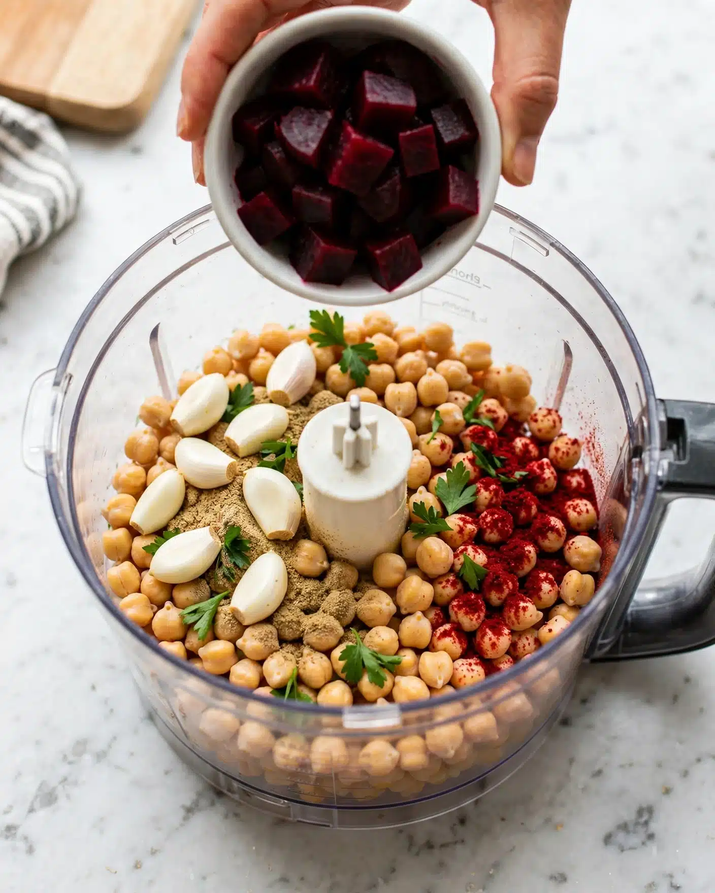Creamy Lemon Beet Hummus Recipe 4 A clear food processor bowl filled with pale yellow chickpeas at the bottom layer, topped with small white garlic cloves, green parsley leaves, light brown ground spices, and sprinkled with red powdered spice unevenly on one side. Above the bowl, a woman's hand is holding a white bowl full of dark red, chunky beet pieces that are about to be added. The background is a white marbled surface with a light wooden cutting board partially visible in the upper left corner. Photo taken with an iphone --ar 4:5 --v 7