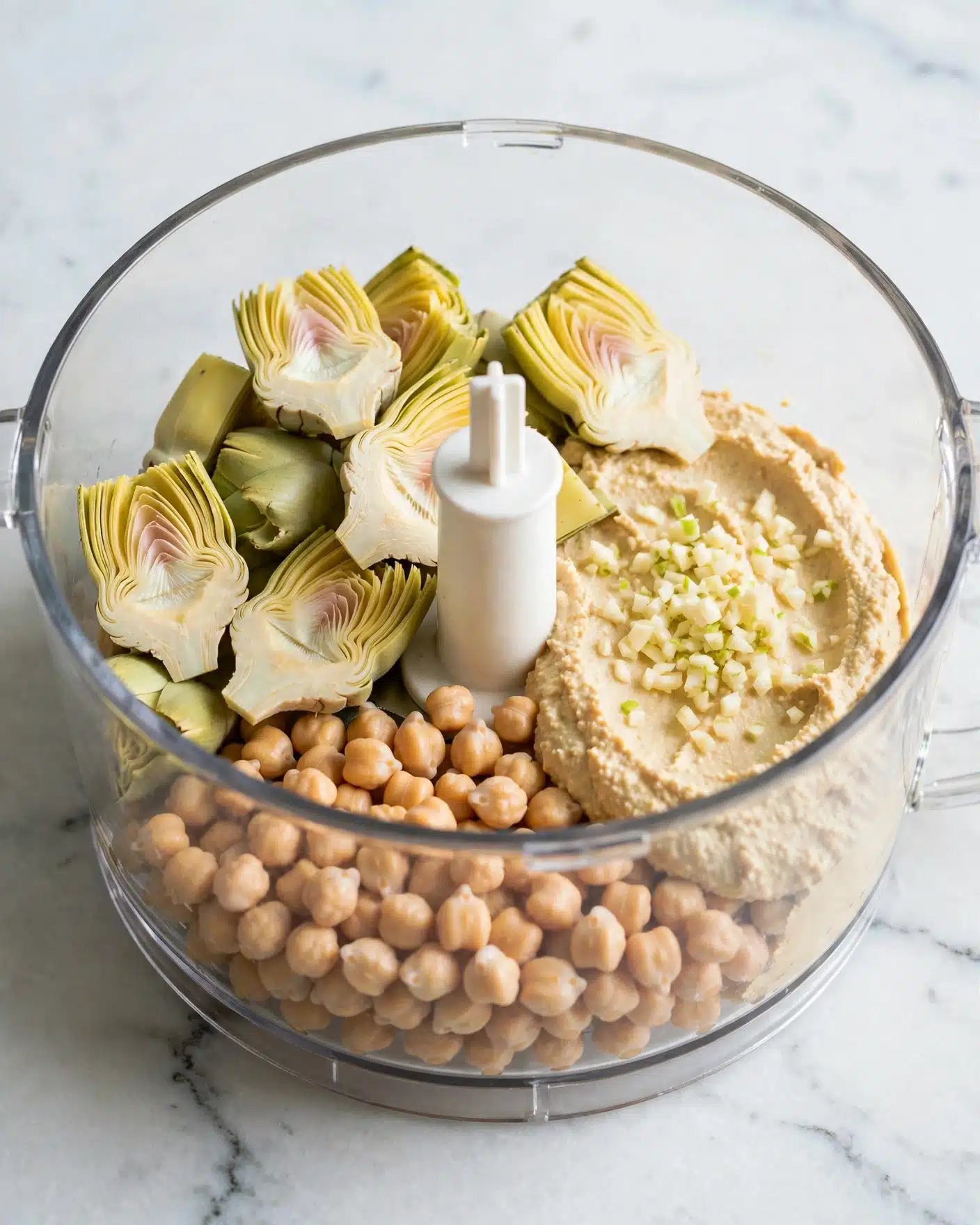 Lemony Artichoke Hummus Recipe 4 A clear round bowl with layers of different ingredients inside placed on a white marbled surface. At the bottom, there are soft beige chickpeas clustered together. Above the chickpeas on the left and spread around the bowl are several artichoke heart quarters with pale yellow and light green colors and a smooth, layered texture. To the right near the artichokes is a thick, creamy light beige hummus-like paste. On the hummus, some finely minced white and light green garlic pieces are sprinkled in the center. The bowl has a central white blade in the middle, part of a food processor. photo taken with an iphone --ar 4:5 --v 7