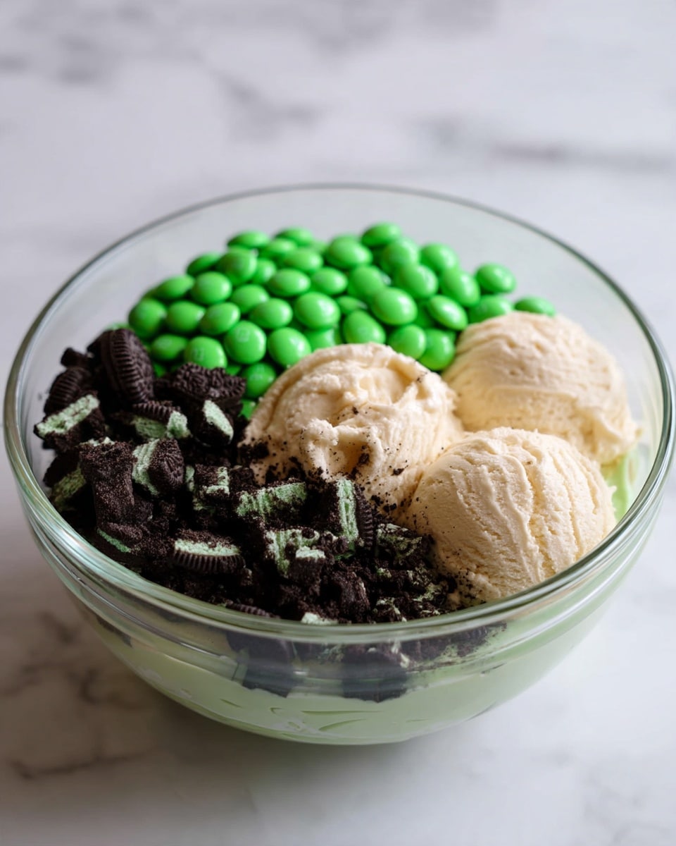 A clear glass bowl sits on a white marbled surface, filled with three main layers of ingredients. On one side, there are bright green candy-coated chocolates piled together smoothly. Next to them is a creamy off-white mix with a soft, thick texture piled in three rounded scoops. The remaining space is filled with crushed mint chocolate sandwich cookies, showing the dark chocolate pieces with light green cream inside spread unevenly over the creamy layer. photo taken with an iphone --ar 4:5 --v 7