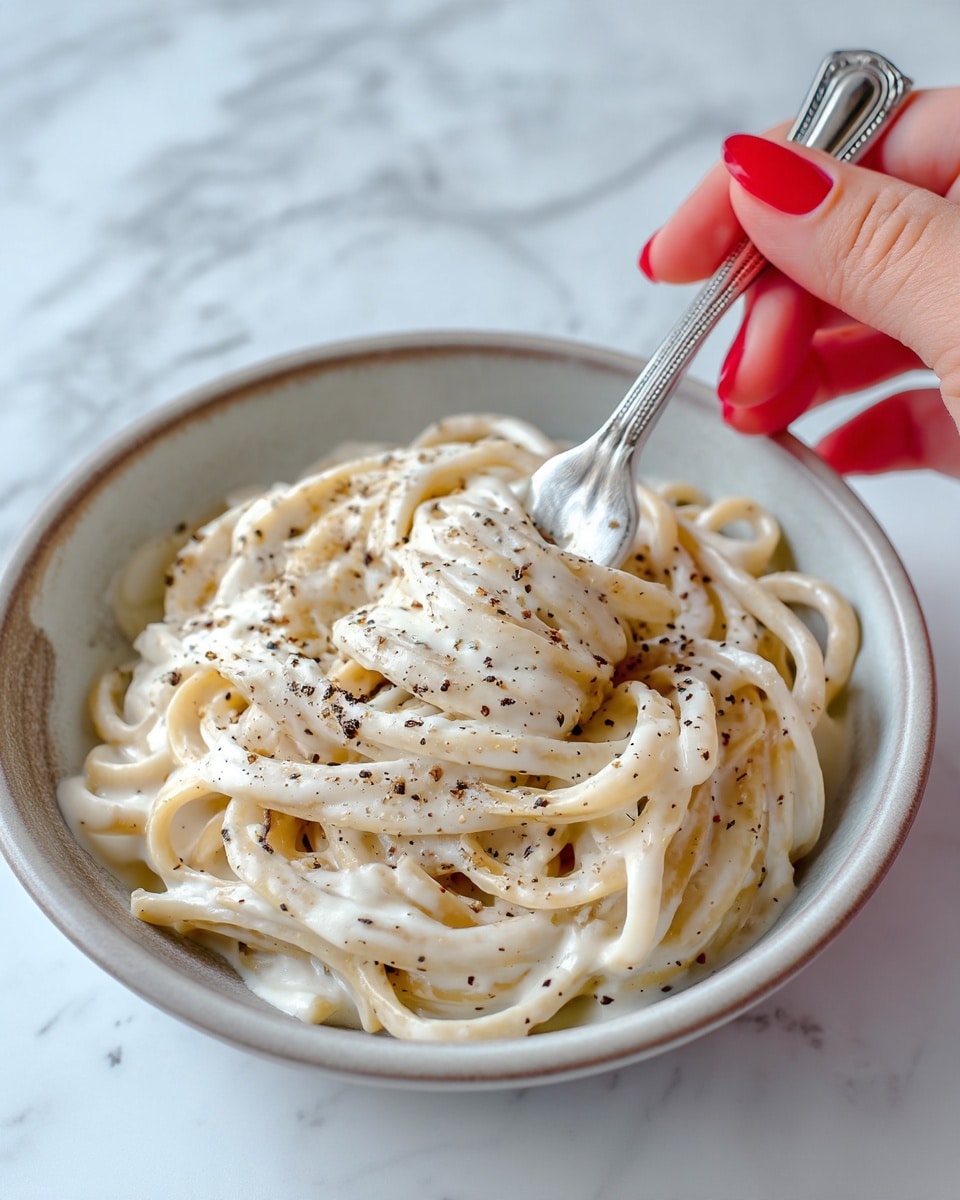 A close-up image of a bowl filled with creamy white pasta coated in a smooth sauce, sprinkled with black pepper. The pasta strands are thick and appear soft, layered slightly unevenly inside a light-colored bowl. A woman's hand with red nail polish holds a silver fork lifting some pasta from the bowl. The background shows a white marbled surface with slight texture and a silver fork lying next to the bowl. Photo taken with an iphone --ar 4:5 --v 7