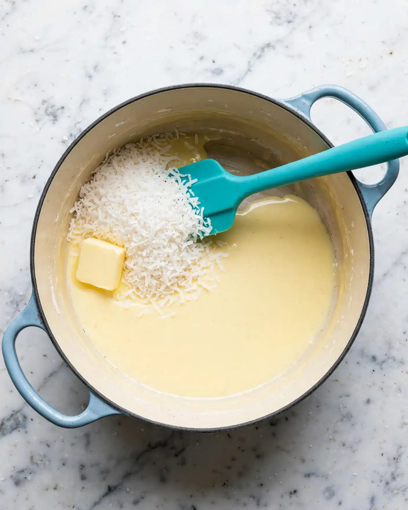 Beijinho (Brazilian Coconut Balls) Recipe 4 A round light blue pot with two handles is shown from above, sitting on a white marbled surface. Inside the pot, there is a layer of light yellow creamy liquid covering half the bottom. On one side of the pot, there is a pile of white shredded coconut resting on the creamy layer. A square piece of pale yellow butter is placed on the creamy liquid near the coconut. A turquoise silicone spatula is resting in the pot, its blade partly under the shredded coconut and its handle extending towards the edge of the pot. Photo taken with an iphone --ar 4:5 --v 7