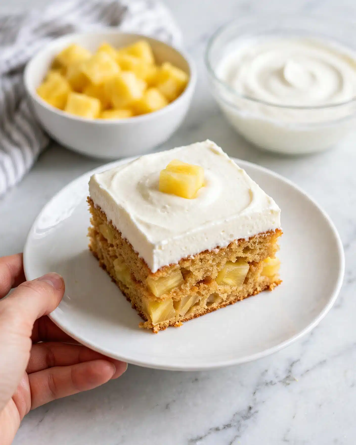 The image shows a square piece of cake on a white plate with a white marbled surface below. The cake has two layers; the bottom layer is a golden-brown color with visible pineapple chunks inside, and the top layer is a thick, smooth white cream spread evenly over the cake. On top of the cream, there is a small yellow piece of pineapple placed in the center. In the background, there is a white bowl filled with more pineapple chunks and a glass bowl with white cream. A woman's hand is holding the plate gently from the side. Photo taken with an iphone --ar 4:5 --v 7