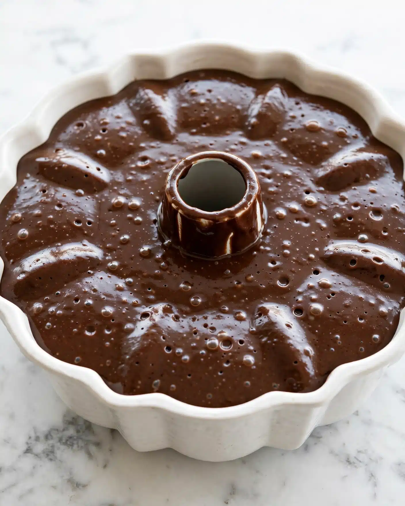Moist Chocolate Bundt Cake with Mini Chocolate Chips and Silky Ganache Recipe 4 A close-up view of a round white Bundt pan filled with dark brown chocolate batter, smooth and glossy in texture with small bubbles on the surface. The batter is evenly spread inside the pan, showing the ridged edges and the central hollow tube of the pan. The pan sits on a white marbled surface, creating a clean and bright background. photo taken with an iphone --ar 4:5 --v 7