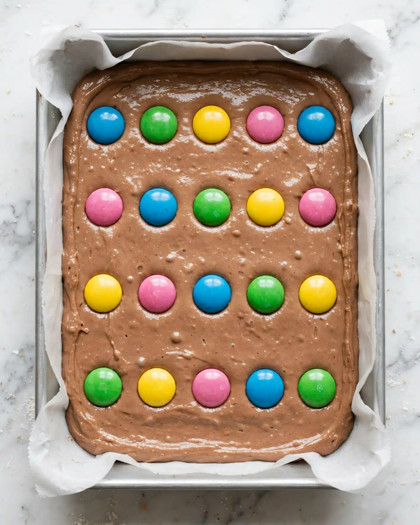 M&M Brownies with Chocolate Frosting Recipe 4 A baking pan lined with white parchment paper holds a single thick layer of smooth, light brown chocolate batter spread evenly across the bottom. On the surface, colorful candy pieces in blue, green, yellow, and pink are placed in neat rows with equal spacing, slightly pressed into the batter but still retaining their round, shiny shape. The background shows a white marbled surface that contrasts softly with the batter. photo taken with an iphone --ar 4:5 --v 7