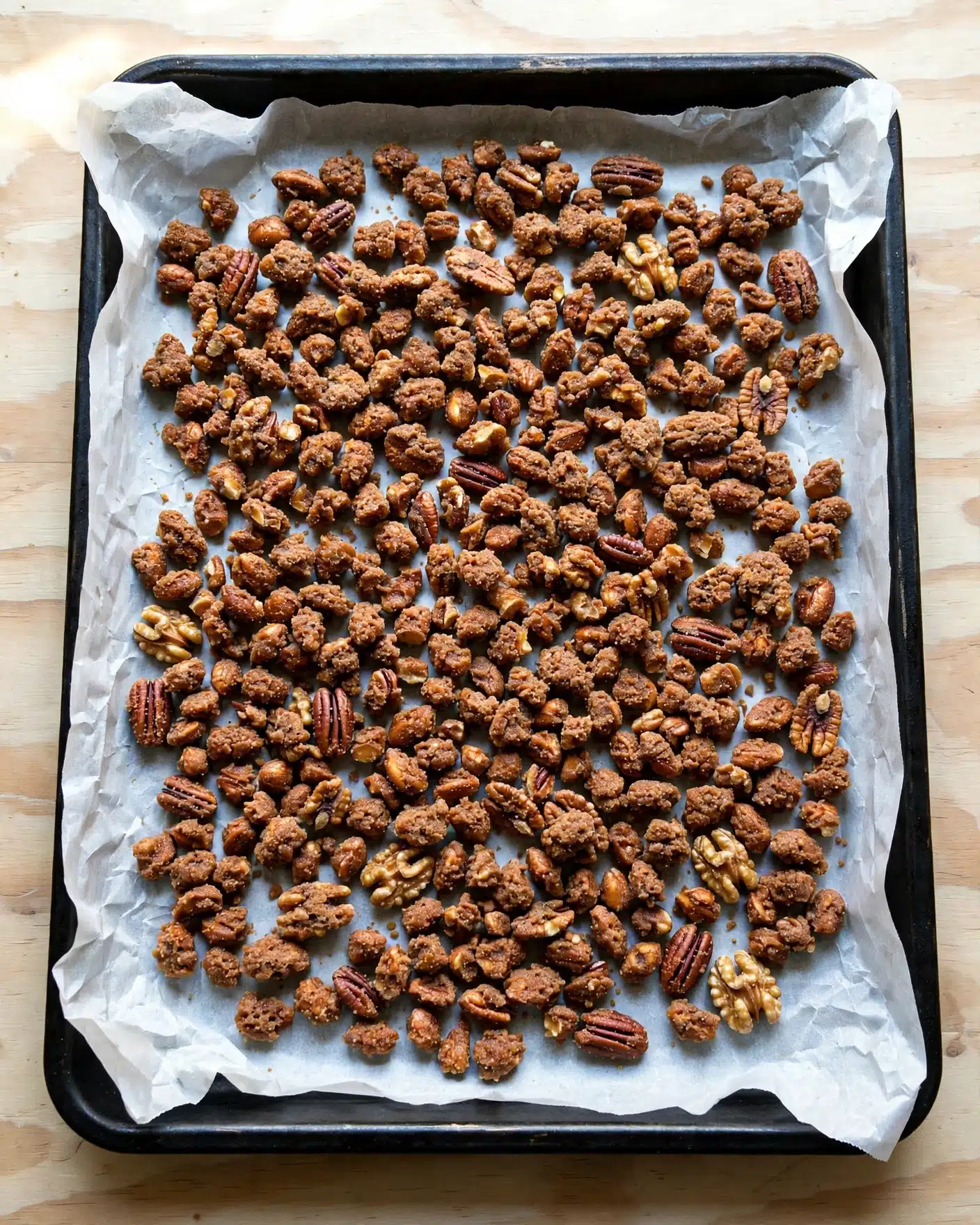 A black rectangular baking tray filled with many uneven pieces of brown candied nuts on white parchment paper. The tray is placed on a wooden surface with a light beige and natural wood grain pattern. The candied nuts have a rough, crunchy texture and are spread out to cover most of the tray's surface. photo taken with an iphone --ar 4:5 --v 7