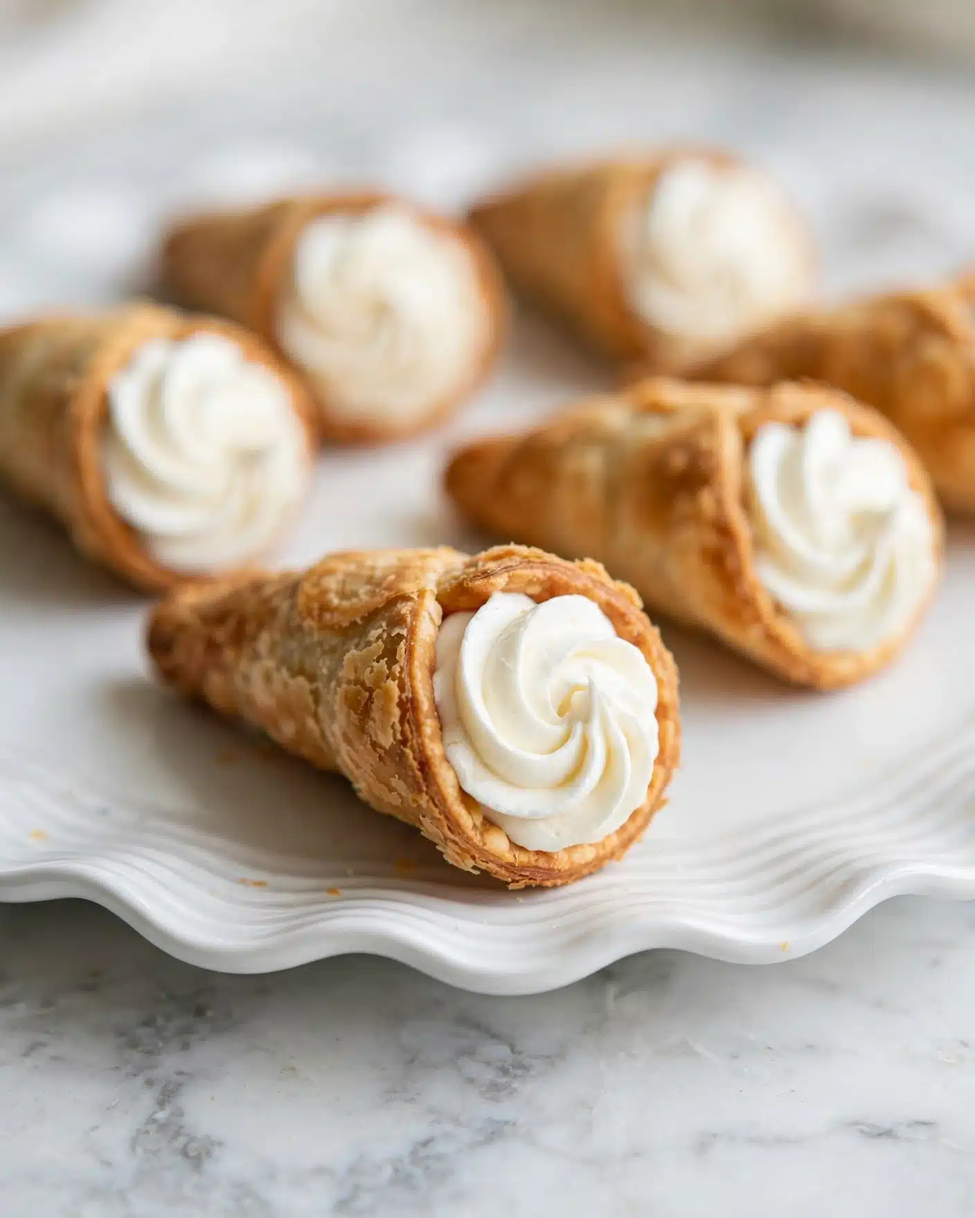 Homemade Cream Horns with Flaky Puff Pastry and Whipped Cream Filling Recipe 5 The image shows several small cream-filled pastry cones placed on a white, wavy-edged plate. Each cone has a golden-brown, crispy outer layer with a textured, slightly flaky surface. The cones are filled with a smooth, white whipped cream, piped in a spiral pattern visible at the wide open end of each cone. The plate is set on a white marbled surface, which creates a clean and bright background for the pastries. The focus is on one cone in the forefront with the rest softly blurred in the background, giving depth to the image. Photo taken with an iphone --ar 4:5 --v 7