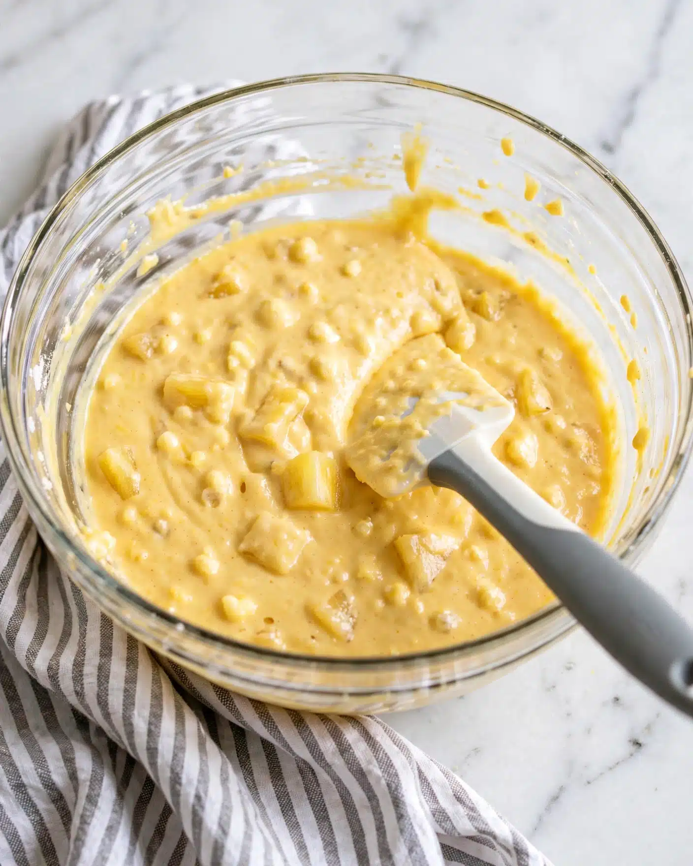 A clear glass bowl filled with a thick yellow batter showing small lumps and a creamy texture. Inside the bowl is a white spatula with a gray handle resting in the batter. The bowl is placed on a white and gray striped cloth on a white marbled surface. The batter fills most of the bowl and looks smooth with some soft pieces inside. photo taken with an iphone --ar 4:5 --v 7
