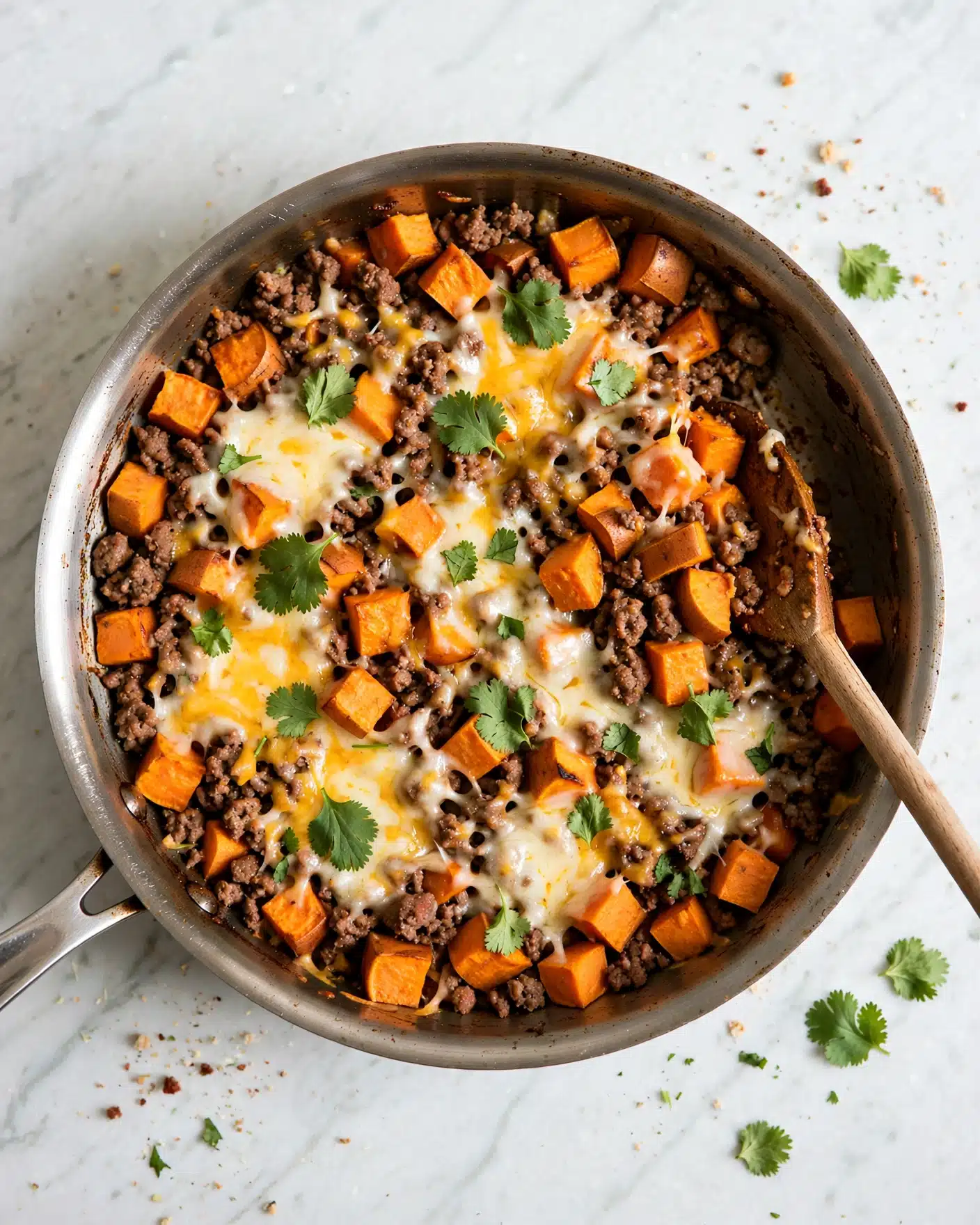 Southwest Ground Beef and Sweet Potato Skillet Recipe 5 The image shows a large metal pan filled with a cooked dish. The dish has a mix of dark brown ground meat and bright orange pieces of cubed sweet potatoes, layered throughout. Melted cheese in creamy white and yellow shades is spread unevenly over the top, blending into the other ingredients. Small green cilantro leaves are scattered across the dish for a fresh touch. A wooden spoon rests inside the pan, partially covered by the food, and the pan is set on a white marbled surface. photo taken with an iphone --ar 4:5 --v 7